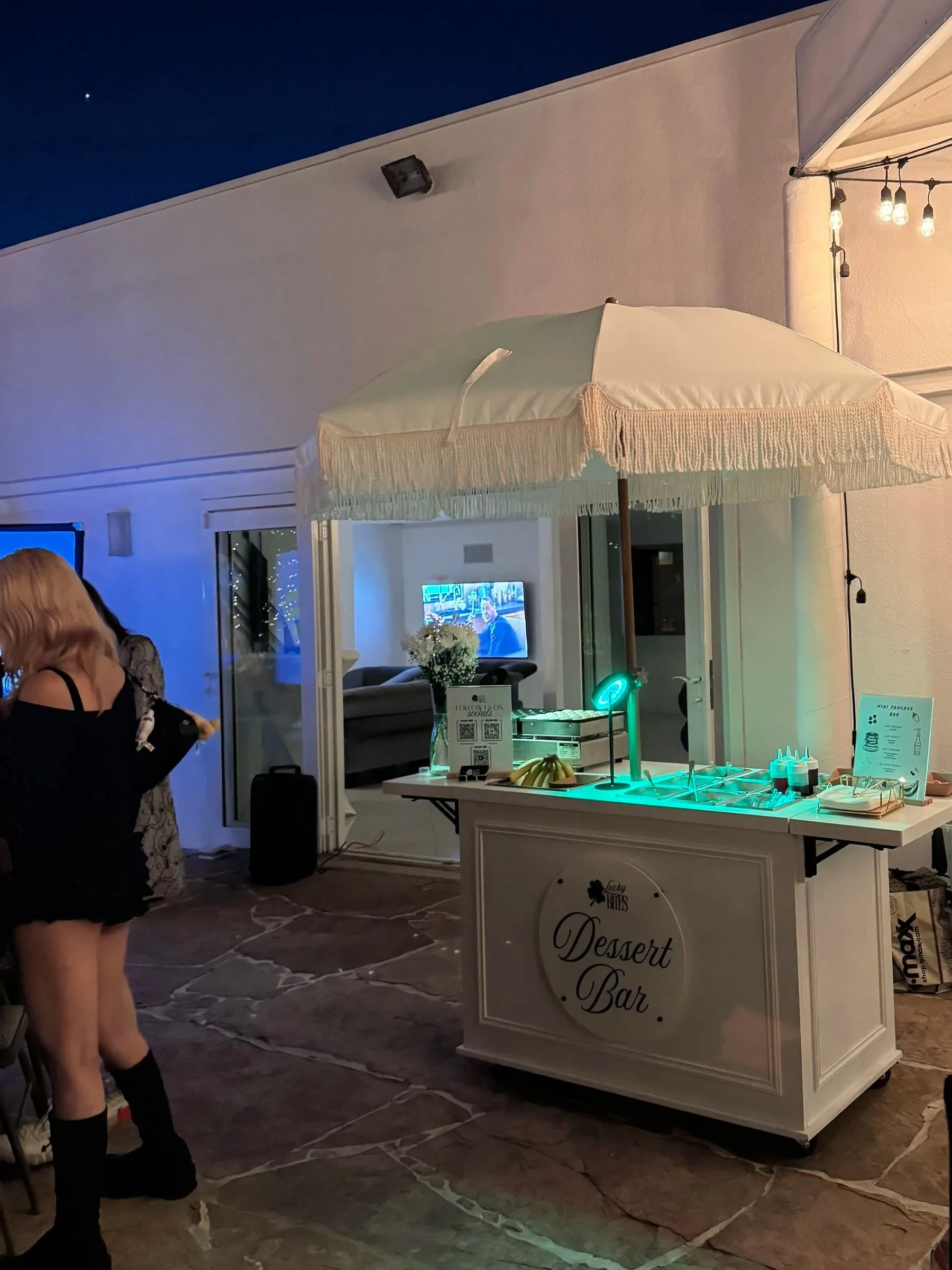 A white dessert bar cart with a fringe umbrella cover illuminated with string lights, positioned on a stone patio at night. In the background, a glass door reveals a TV, a woman with blonde hair, and balloons, indicating a celebration setting.