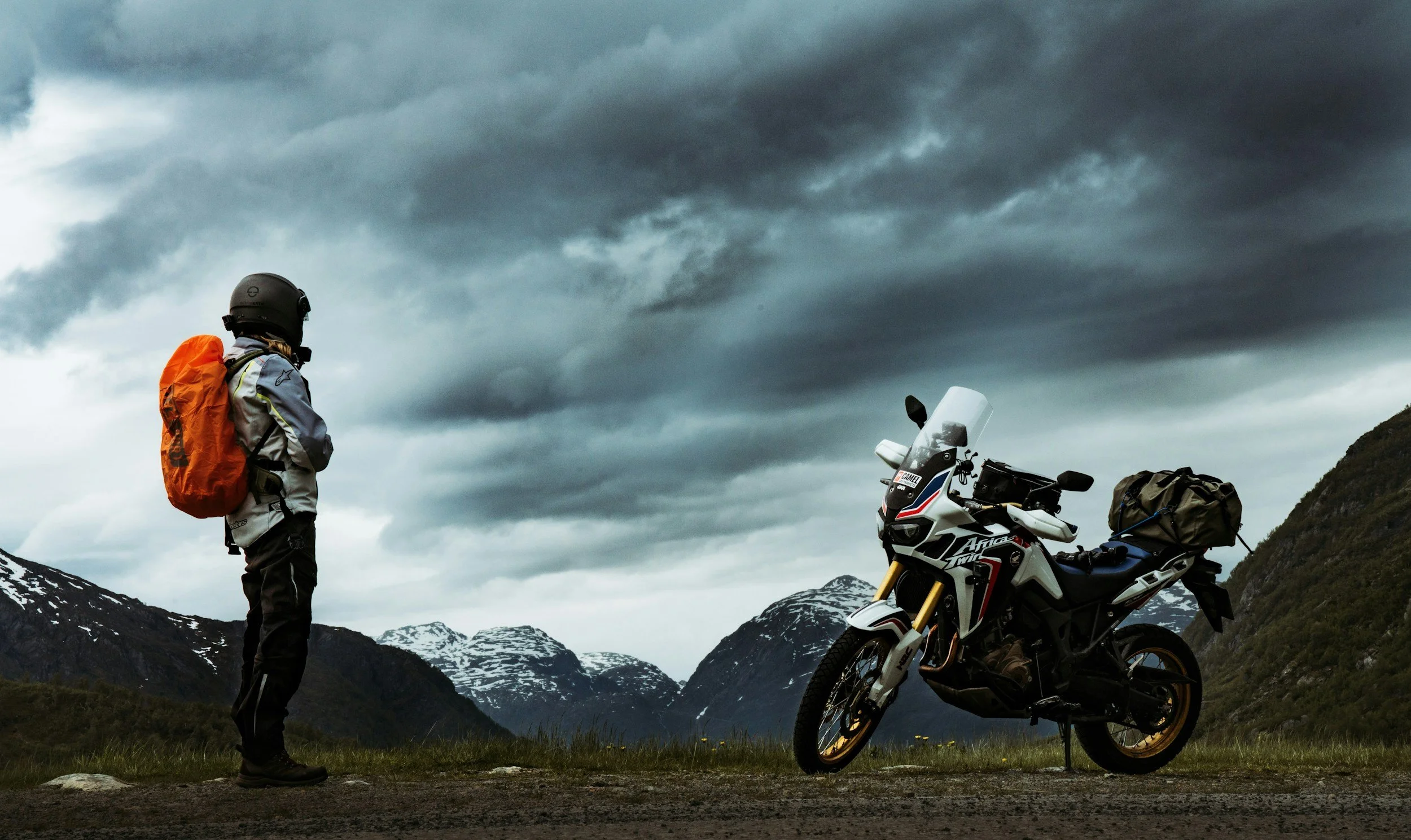 Adventure motorcycle and rider at an off-road campsite with mountains in the distance under a stormy sky.