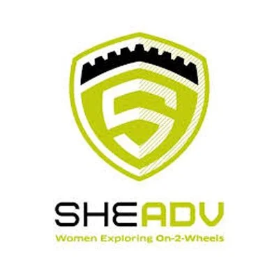 Logo for SHE ADV, a women's group supporting adventure motorcycle riding for women, featuring a shield with a stylized letter S and a crown-like top.