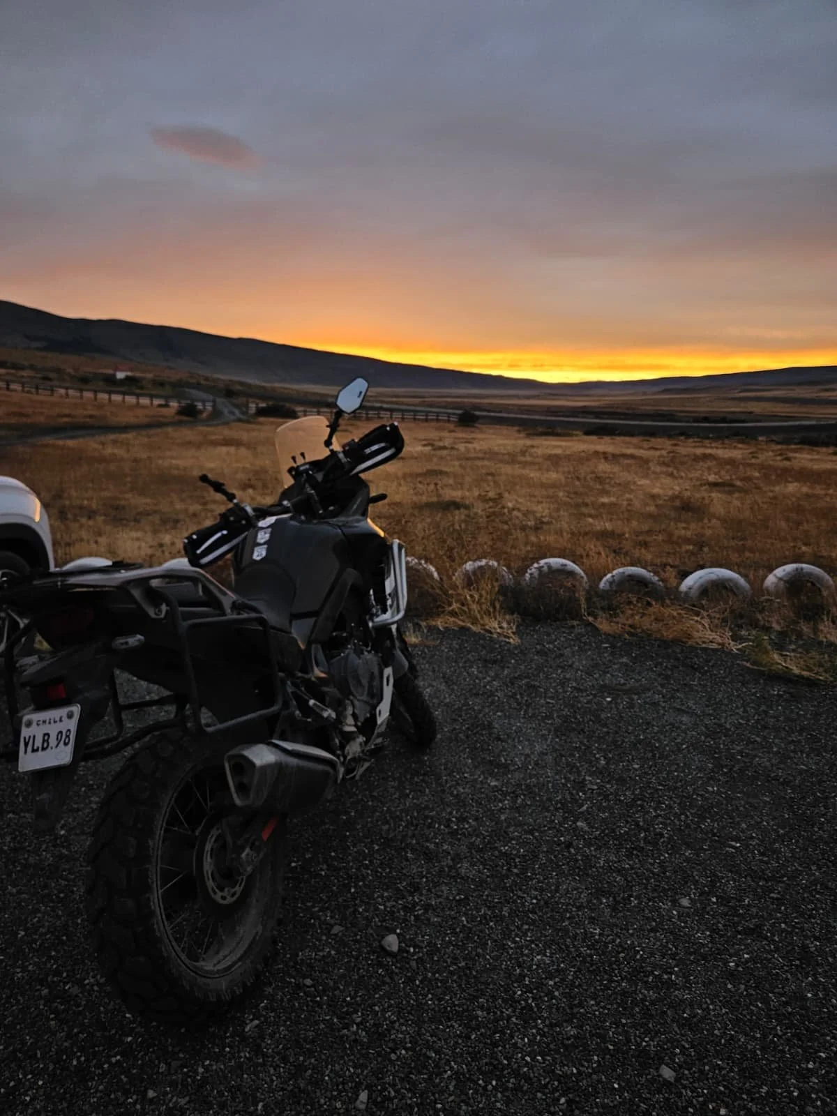 ADV motorcycle parked on gravel facing the dramatic sunset between the distant hills.
