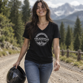 A woman with long dark hair smiling and walking outdoors on a dirt road with trees and mountains in the background, holding a motorcycle helmet.