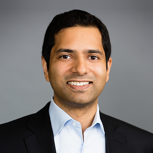 Professional headshot of a man in a black suit and white shirt smiling at the camera with a gray background.