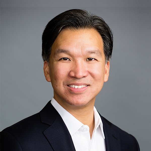 Professional headshot of a smiling man in a black suit and white shirt against a gray background.