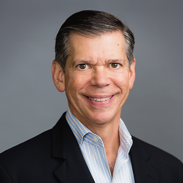 Portrait of a smiling middle-aged man with dark hair, wearing a dark suit and light blue striped shirt, against a gray background.