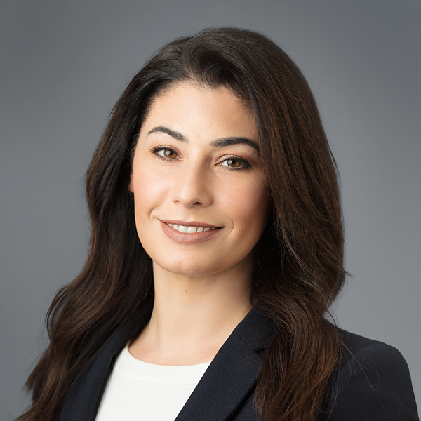 Professional woman with long, dark hair, smiling, wearing a dark blazer and white top against a gray background.