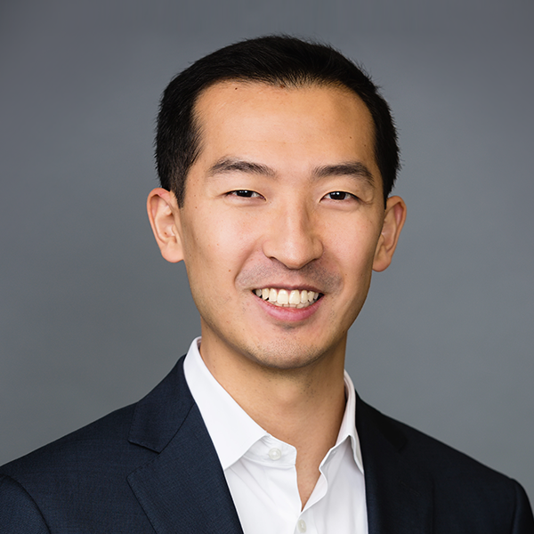 A man with short black hair smiling, dressed in a white shirt and dark blazer against a gray background.