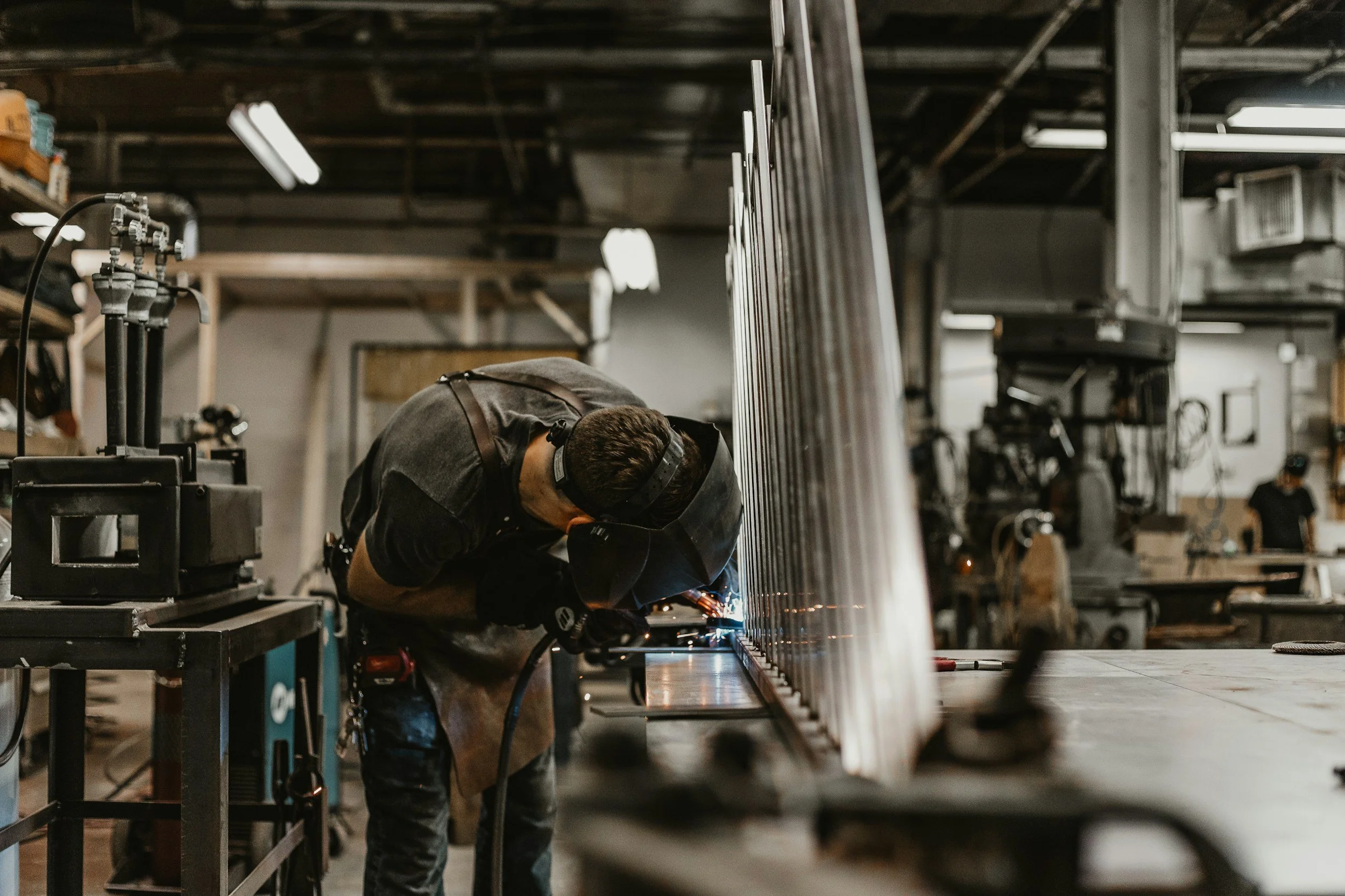 A person welding metal panels in a workshop filled with equipment and lighting fixtures.