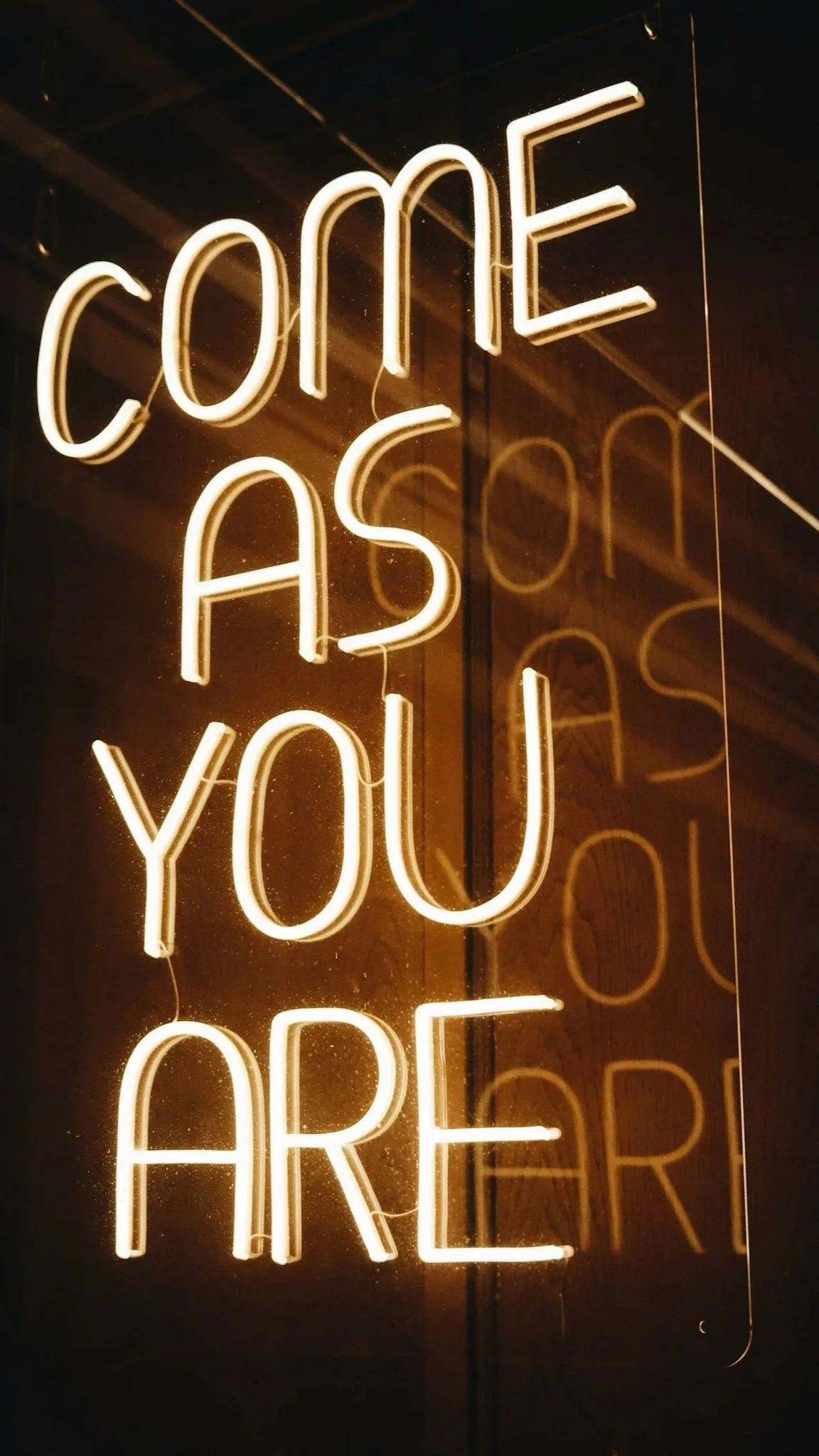 Neon sign with the words "Come As You Are" illuminated in warm white light, reflected on a glass surface.