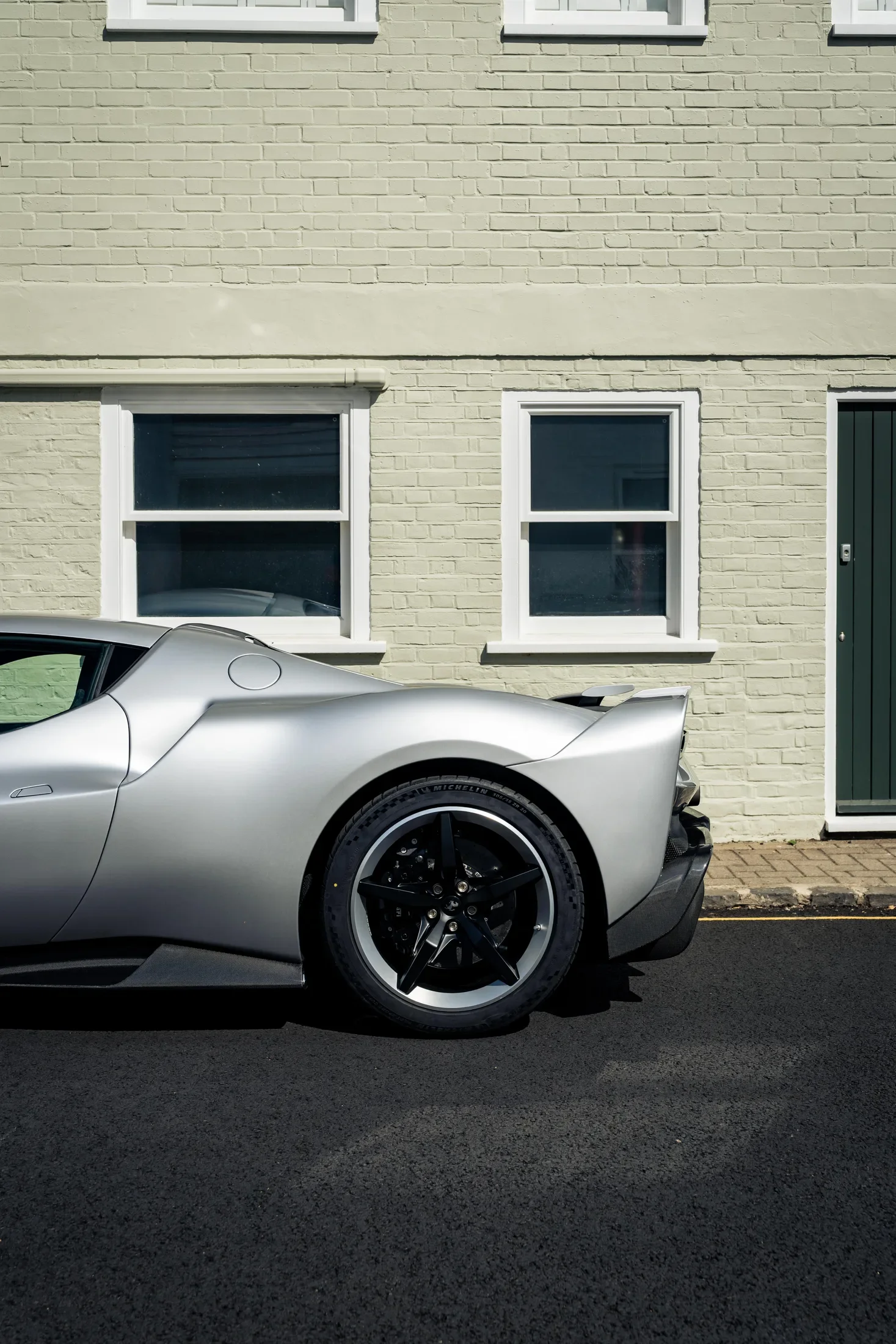 Ferrari 296 Speciale in Argento Nürburgring — Rear quarter beside a pale brick house — AKC Asset Management.