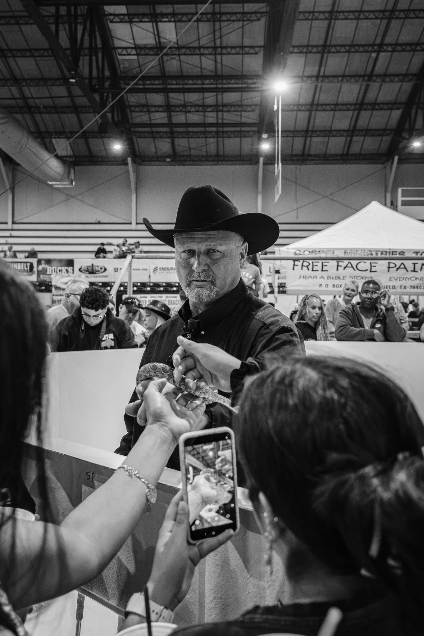 A man wearing a cowboy hat is holding a snake out for the public to touch at a public event in an indoor arena. Fans are taking photos and holding out their phones to capture the moment. A banner in the background advertises free face painting.