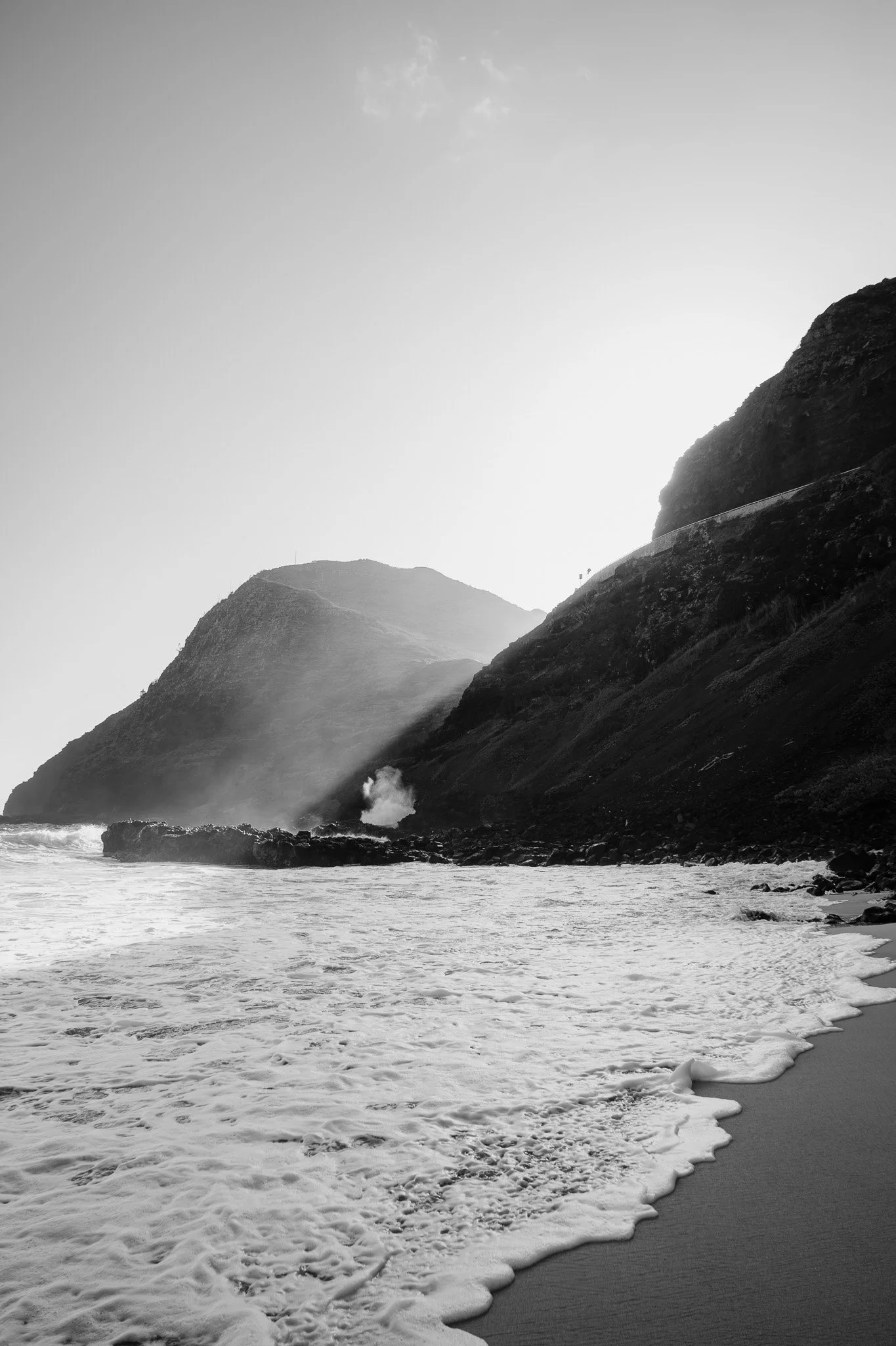 Black and white photograph of a rocky coastline with mountains, ocean waves crashing on the rocks, and clouds in the sky.