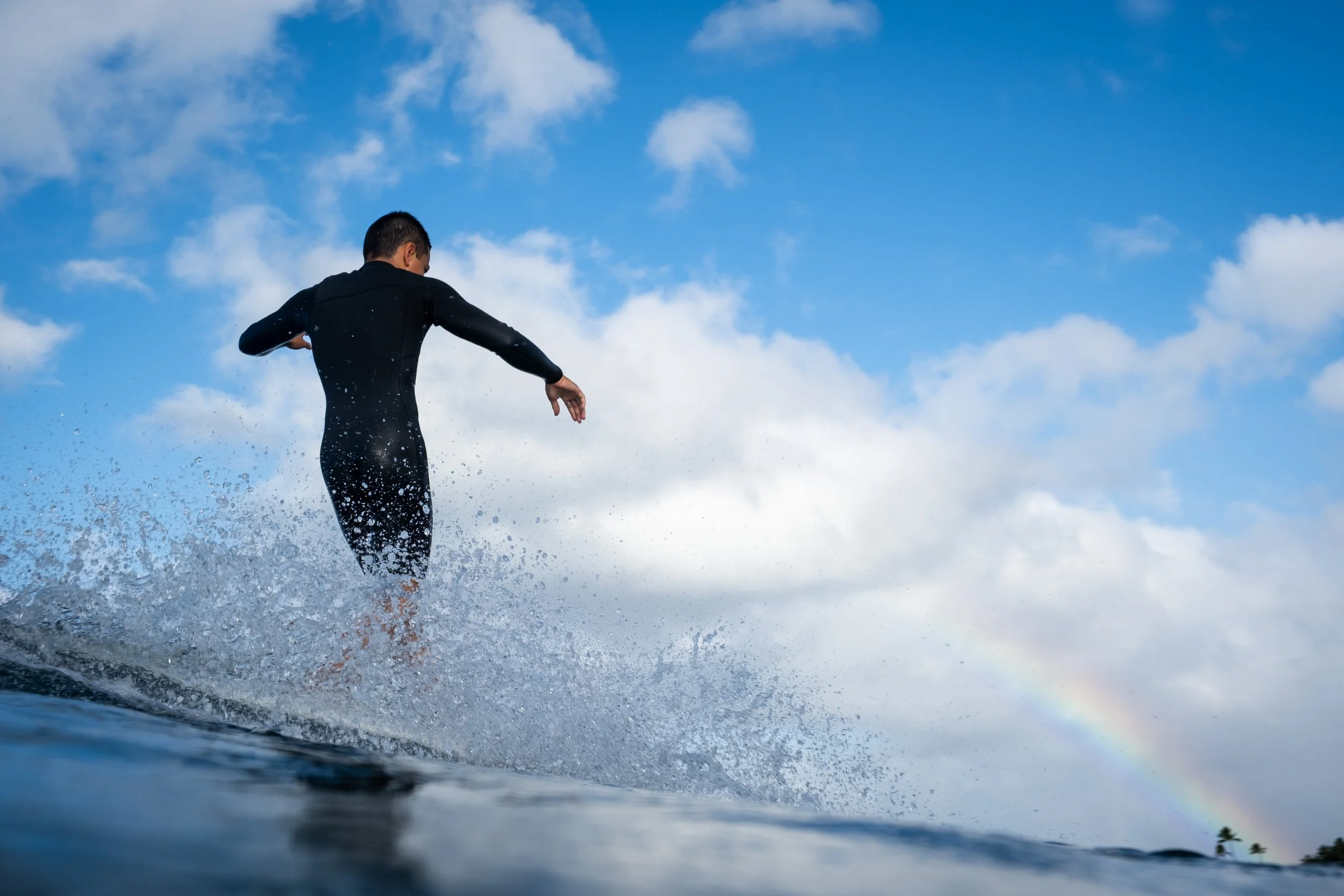 A person wearing a wetsuit standing on a surfboard in the water, with a partly cloudy sky above and rainbow visible in the corner.