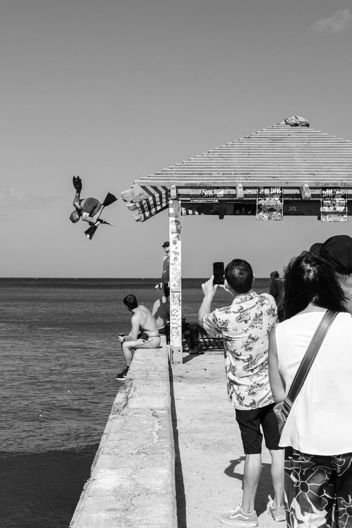 People at seaside watching a person perform a stunt mid-air, jumping from the roof structure into the ocean, in black and white.