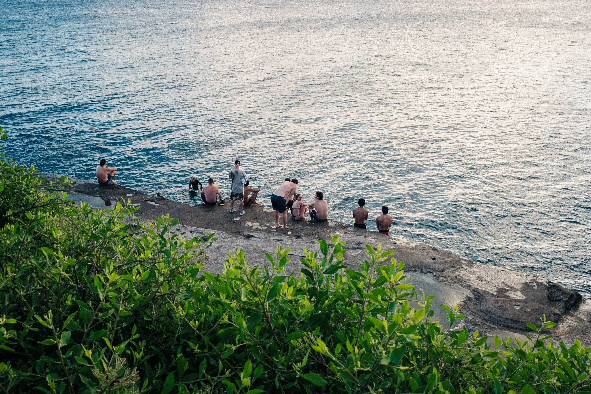People sitting and standing on a rocky shoreline next to a body of water with gentle ripples, surrounded by green vegetation.