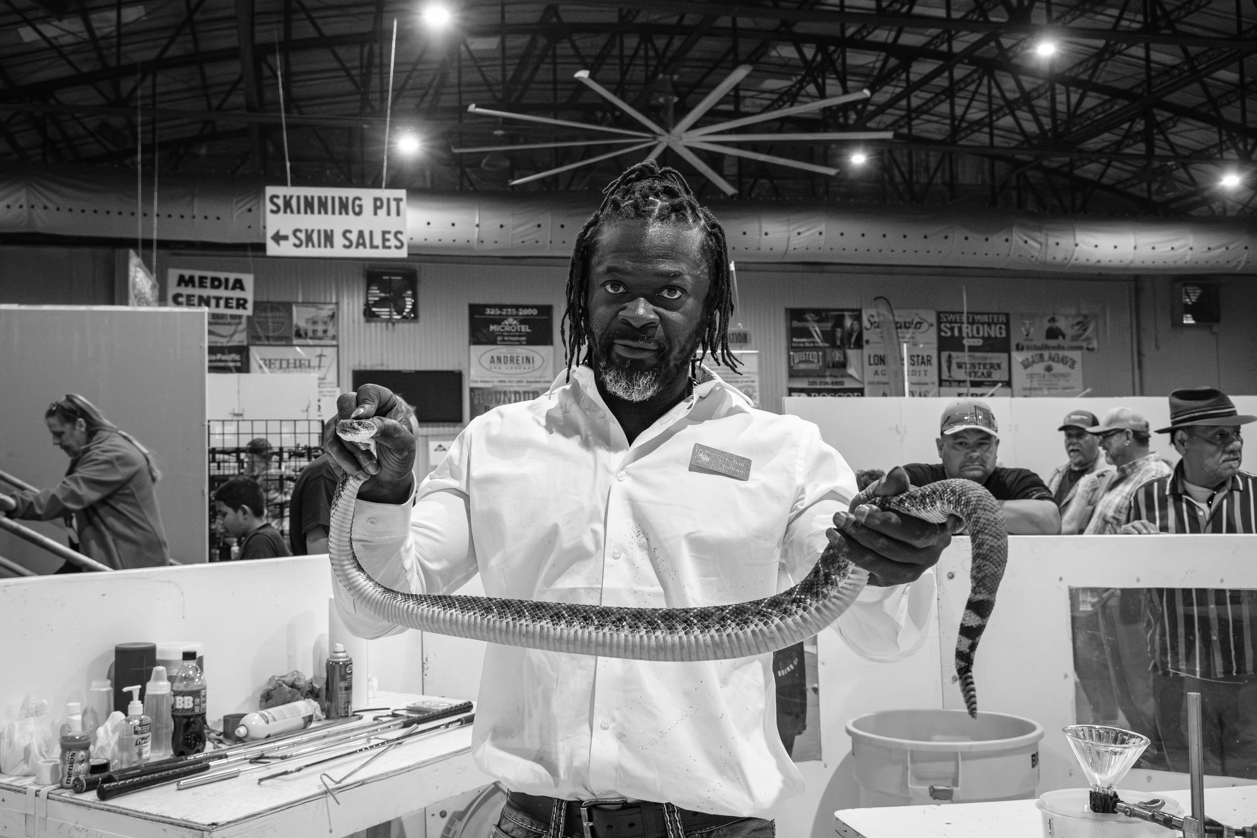 A man with dreadlocks and a grey beard holding a snake at an indoor event with spectators and signs in the background.