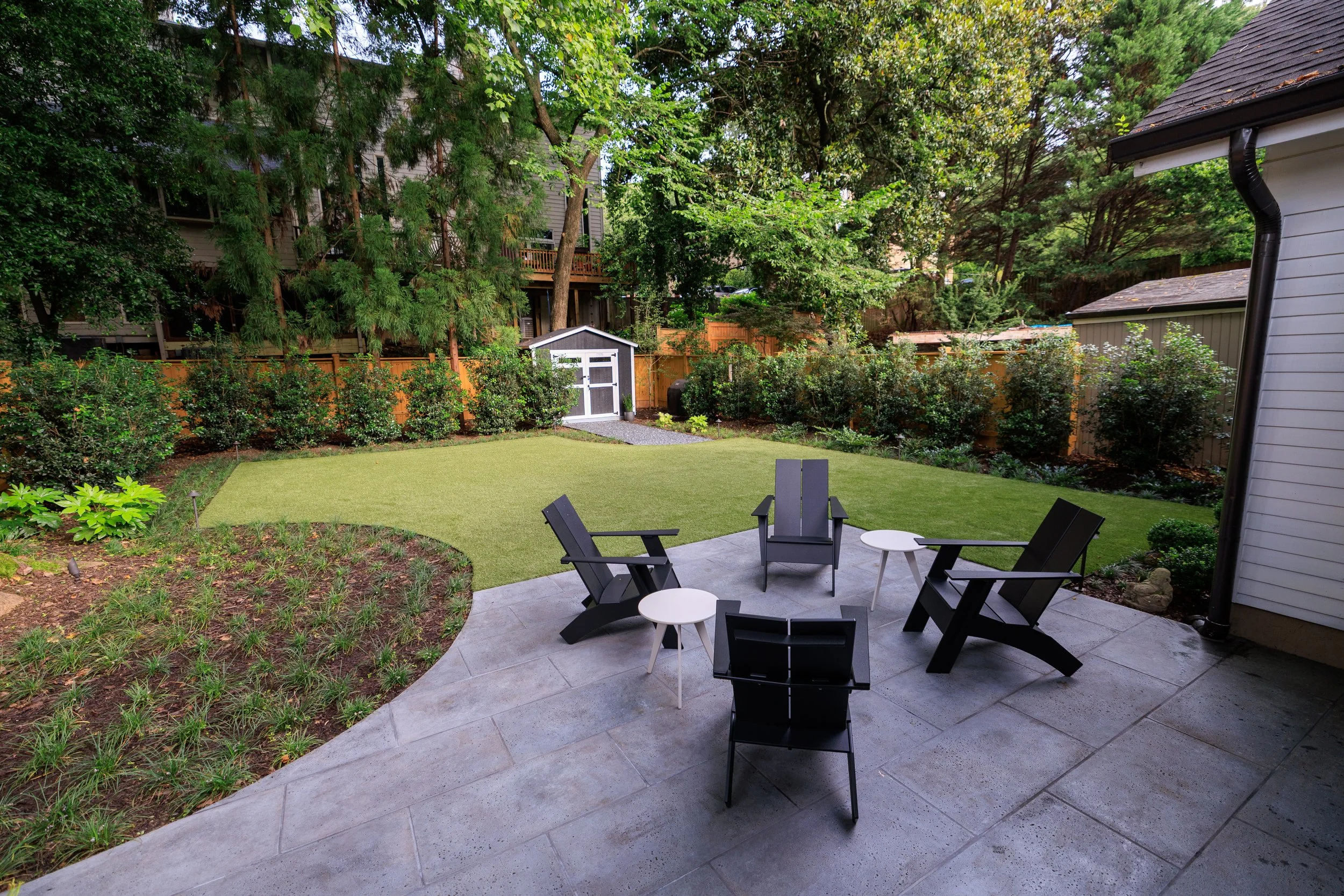 A backyard patio with black chairs and white side tables, surrounded by lush green bushes and trees, with a small shed and house in the background.
