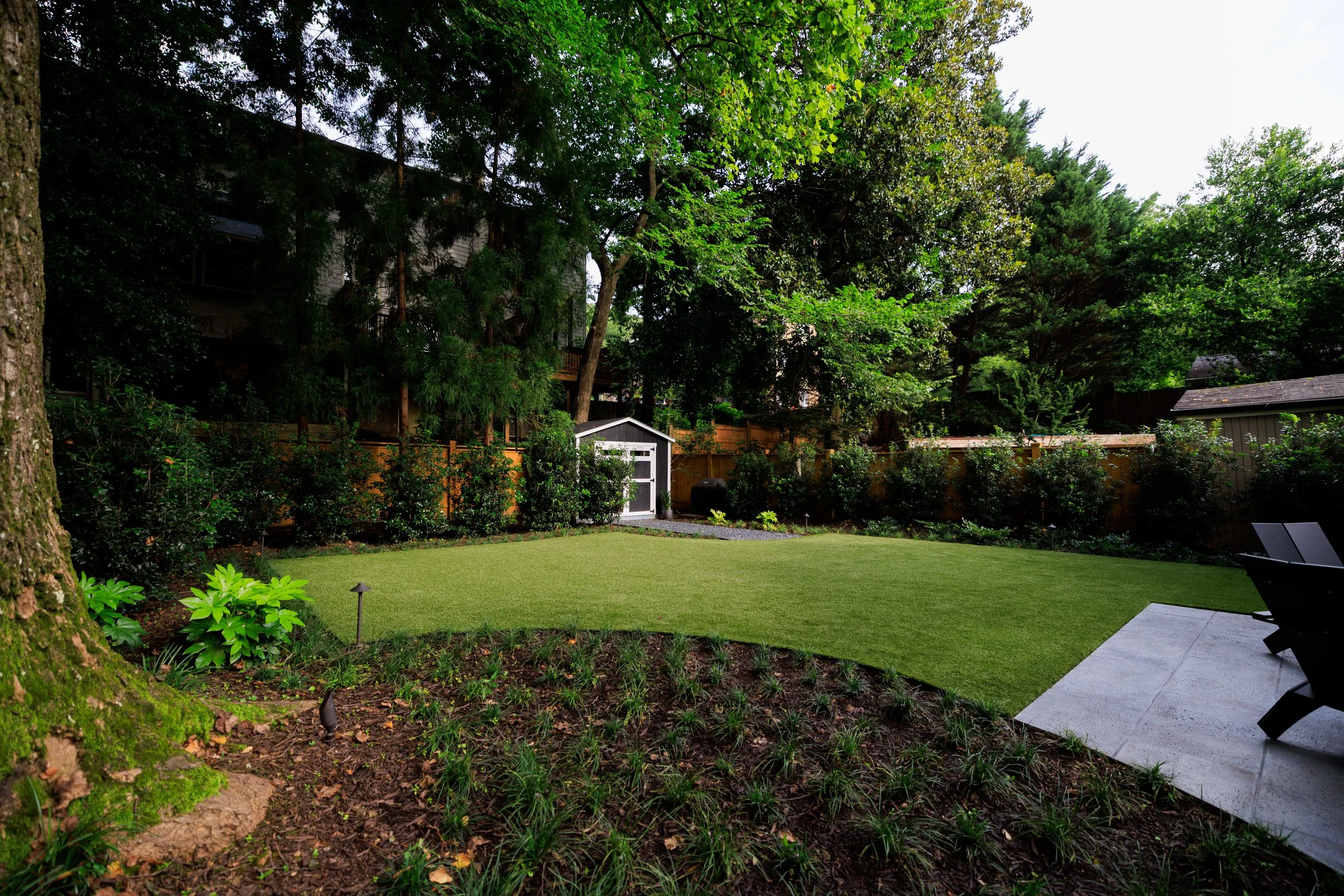 A lush backyard garden with a well-maintained lawn, surrounded by trees and shrubs, a small white garden shed, and outdoor seating.