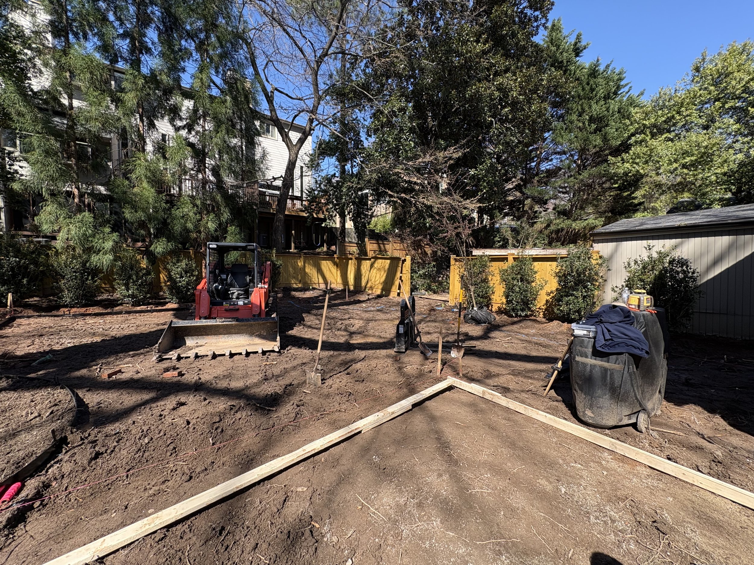 A backyard under construction with bare soil, wooden frames, a small red mini excavator, and trees surrounding the area.
