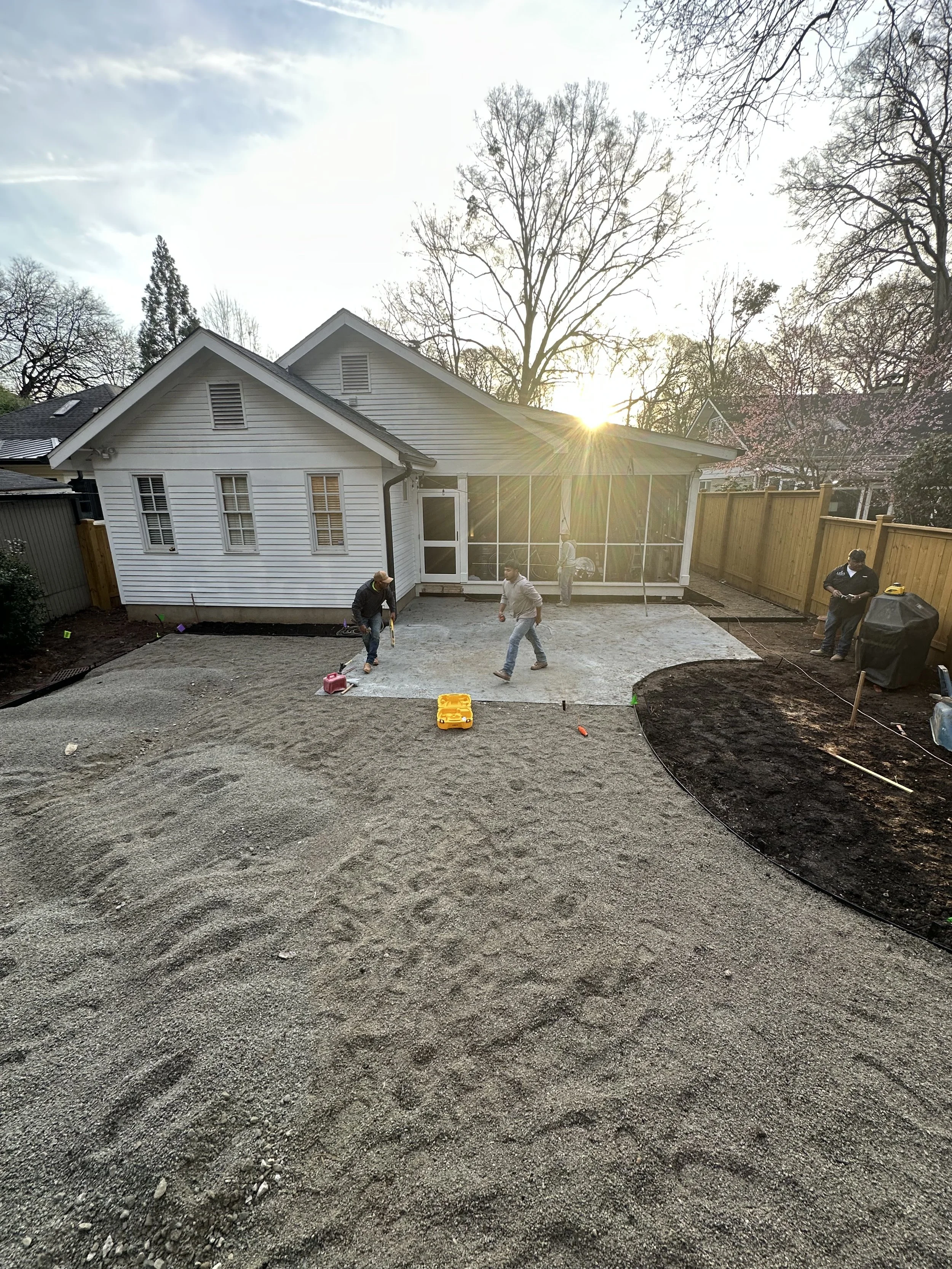 People working on a backyard renovation, preparing the ground for new paving or landscaping, with a white house and trees in the background at sunset.