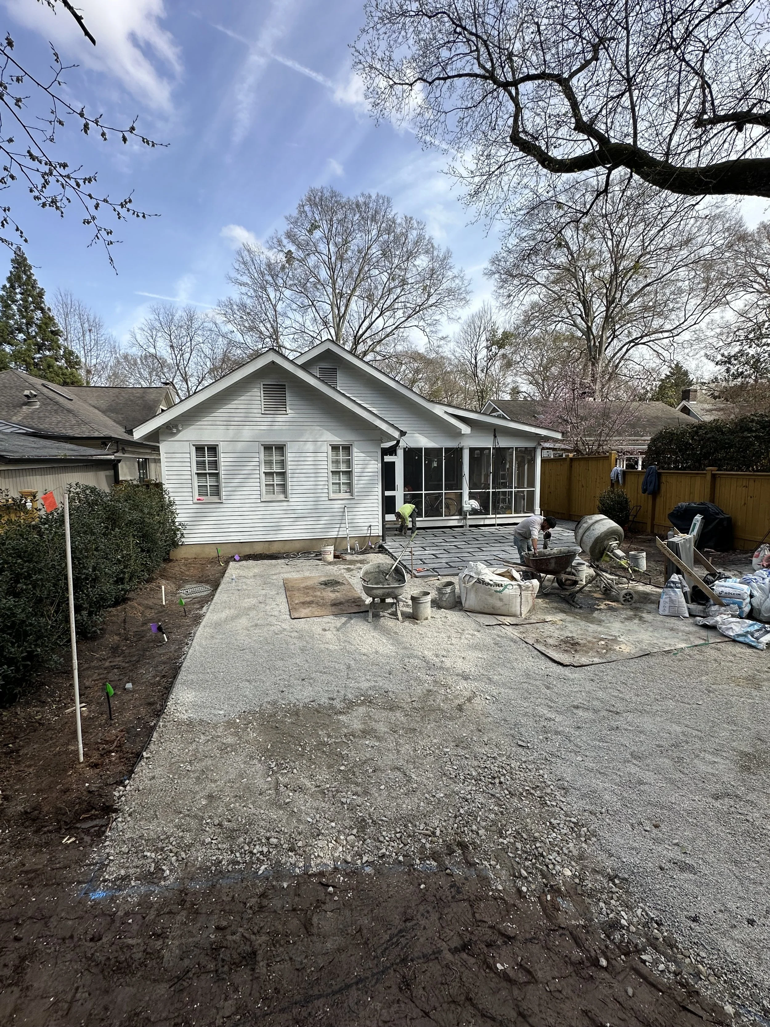 Backyard with a white house, construction workers, and a partially completed concrete patio, with trees in the background and a wooden fence on the right.
