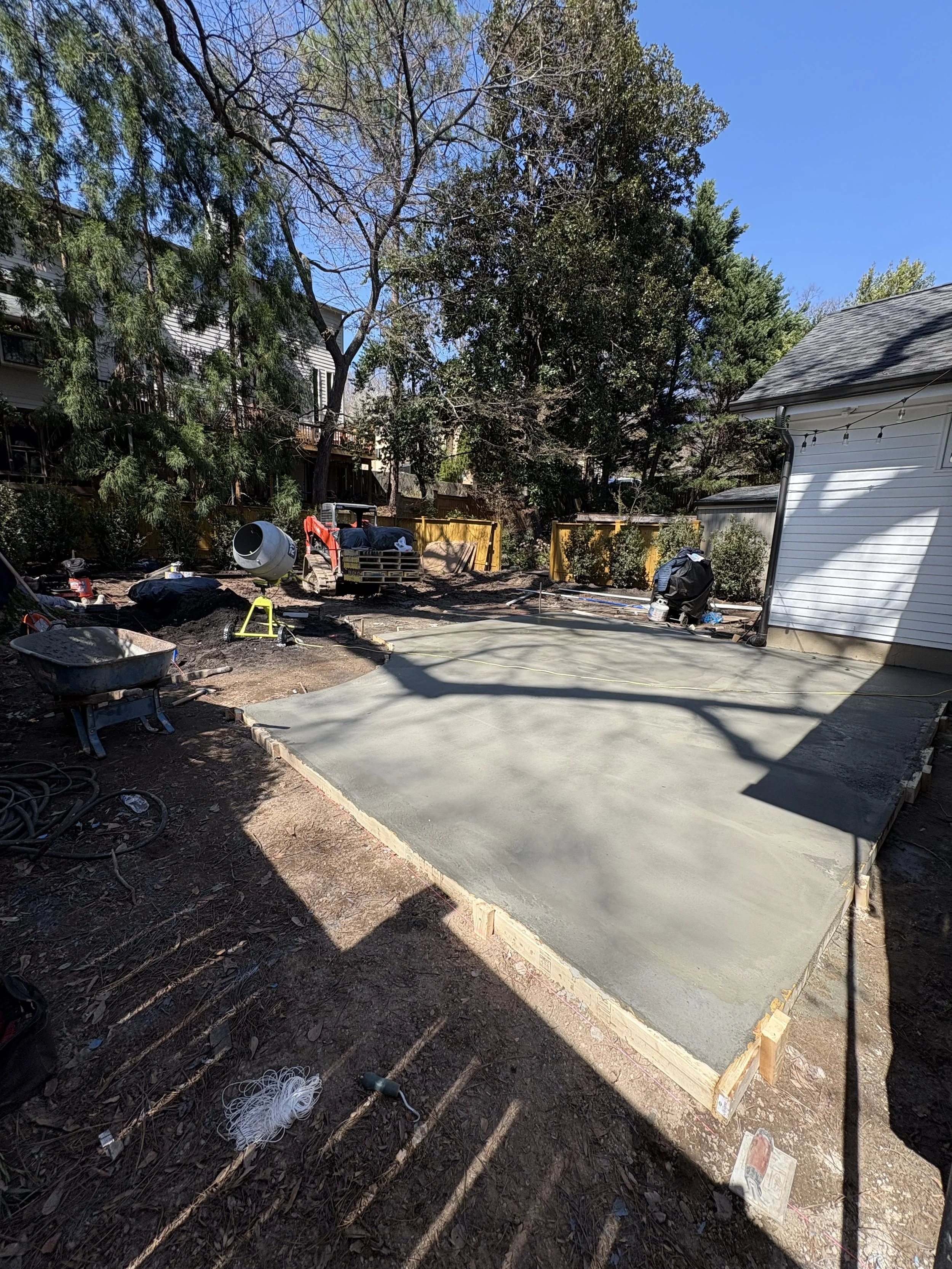 Backyard scene with new concrete slab, construction tools, scattered wires, wheelbarrow, and surrounding trees and houses.