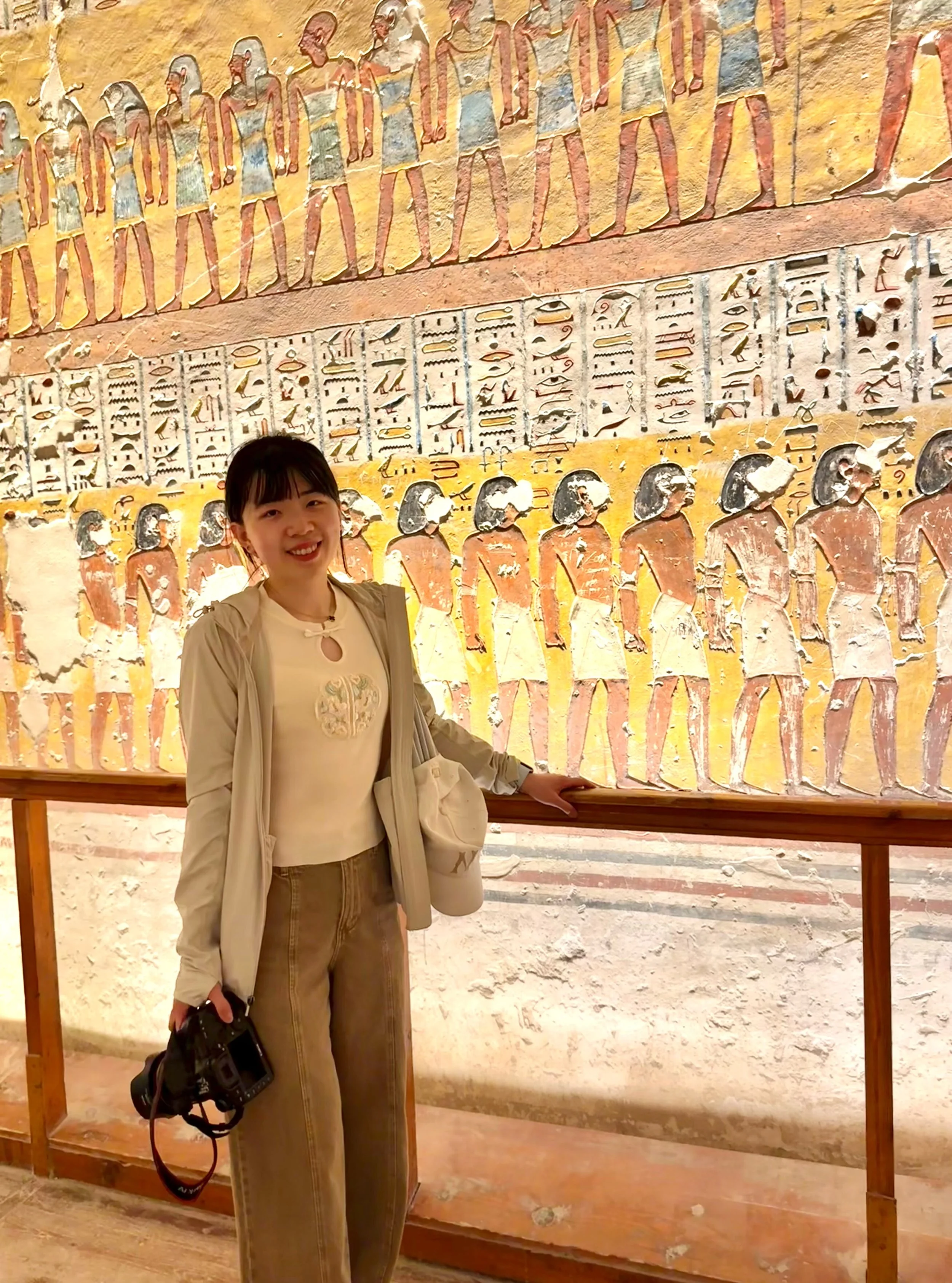 A woman stands in front of an ancient Egyptian wall painting with hieroglyphics and figures. She is smiling, holding a camera, and wearing a beige jacket and pants.
