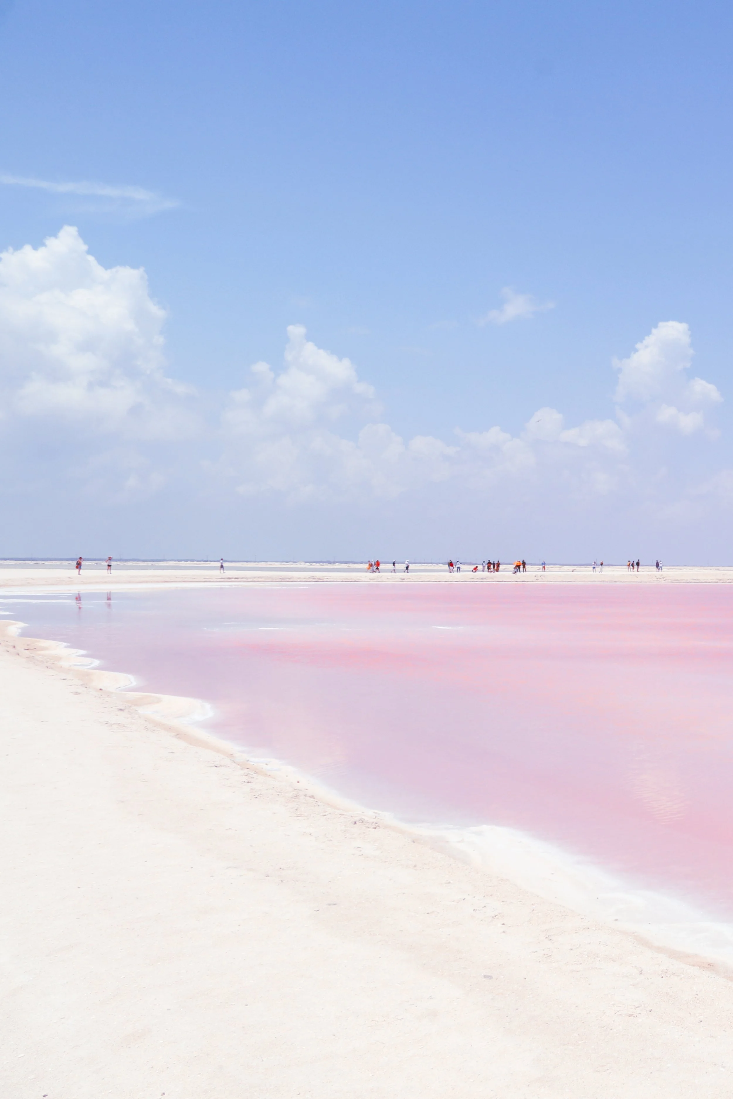People walking and relaxing along the shore of a pink salt lake under a blue sky with clouds.
