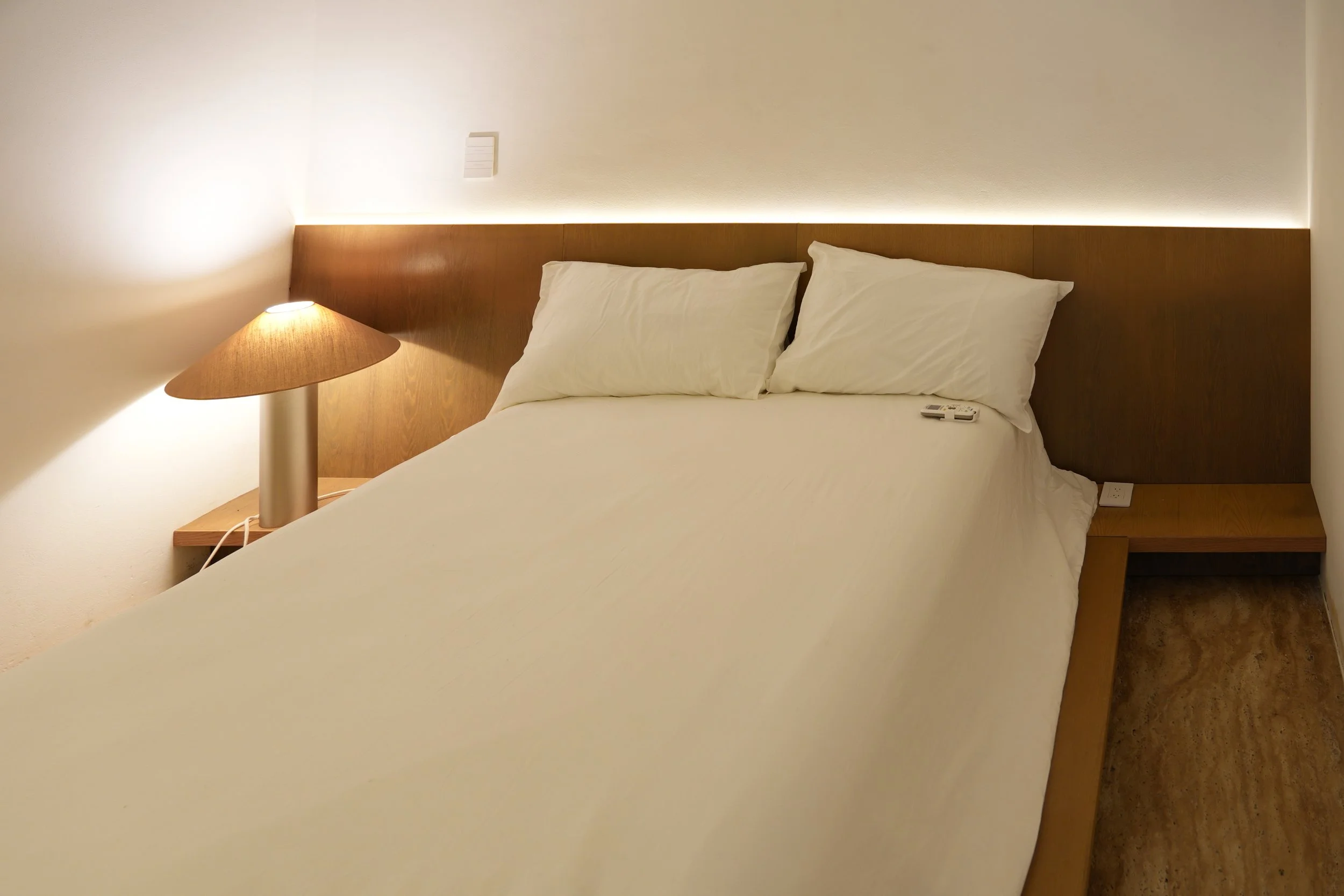 A neatly made bed with white sheets and two white pillows, wooden headboard, bedside lamp, and a remote control on the bed, in a minimalist hotel room.