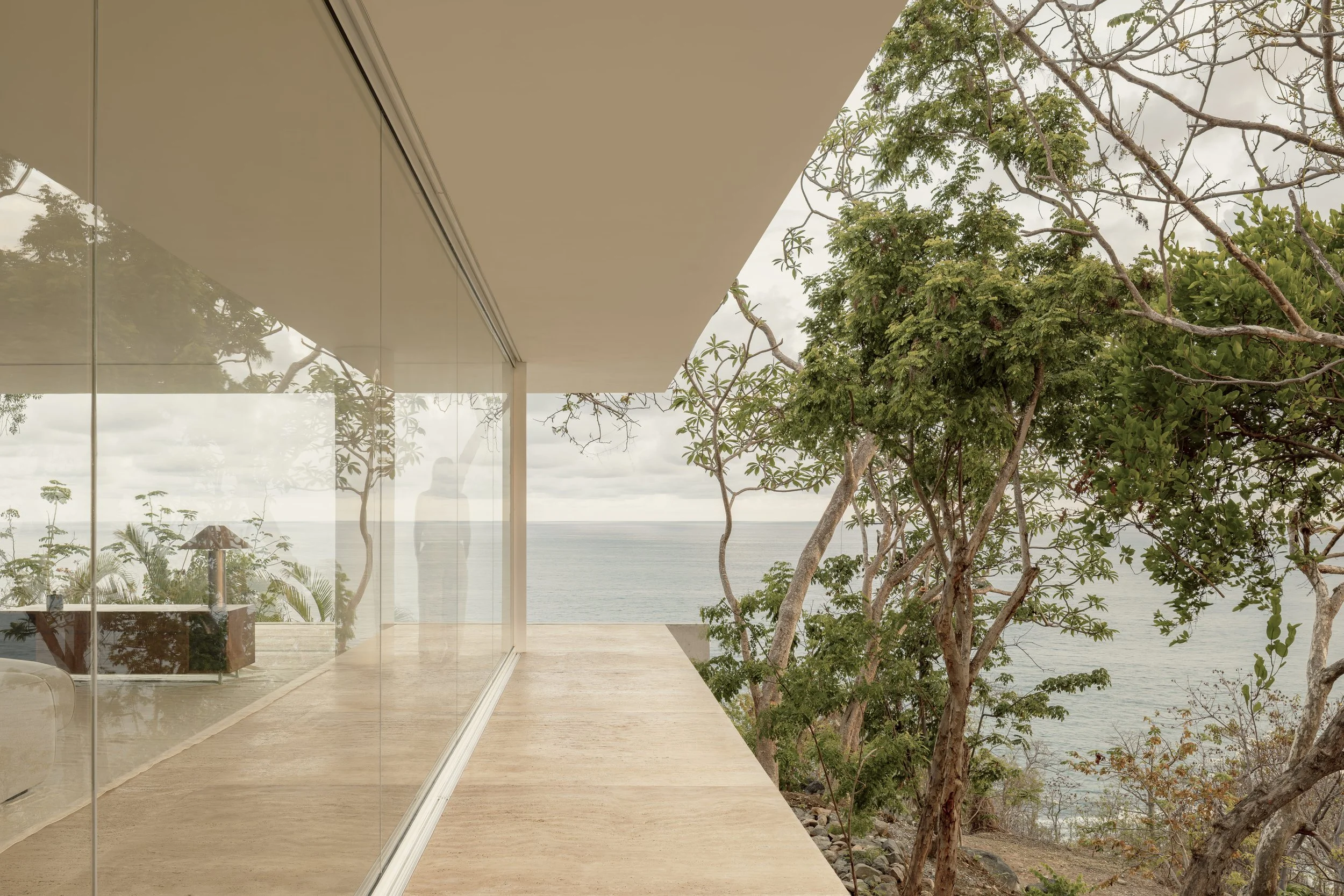 Modern house with glass walls and a wooden deck, overlooking trees and an ocean view during daytime.