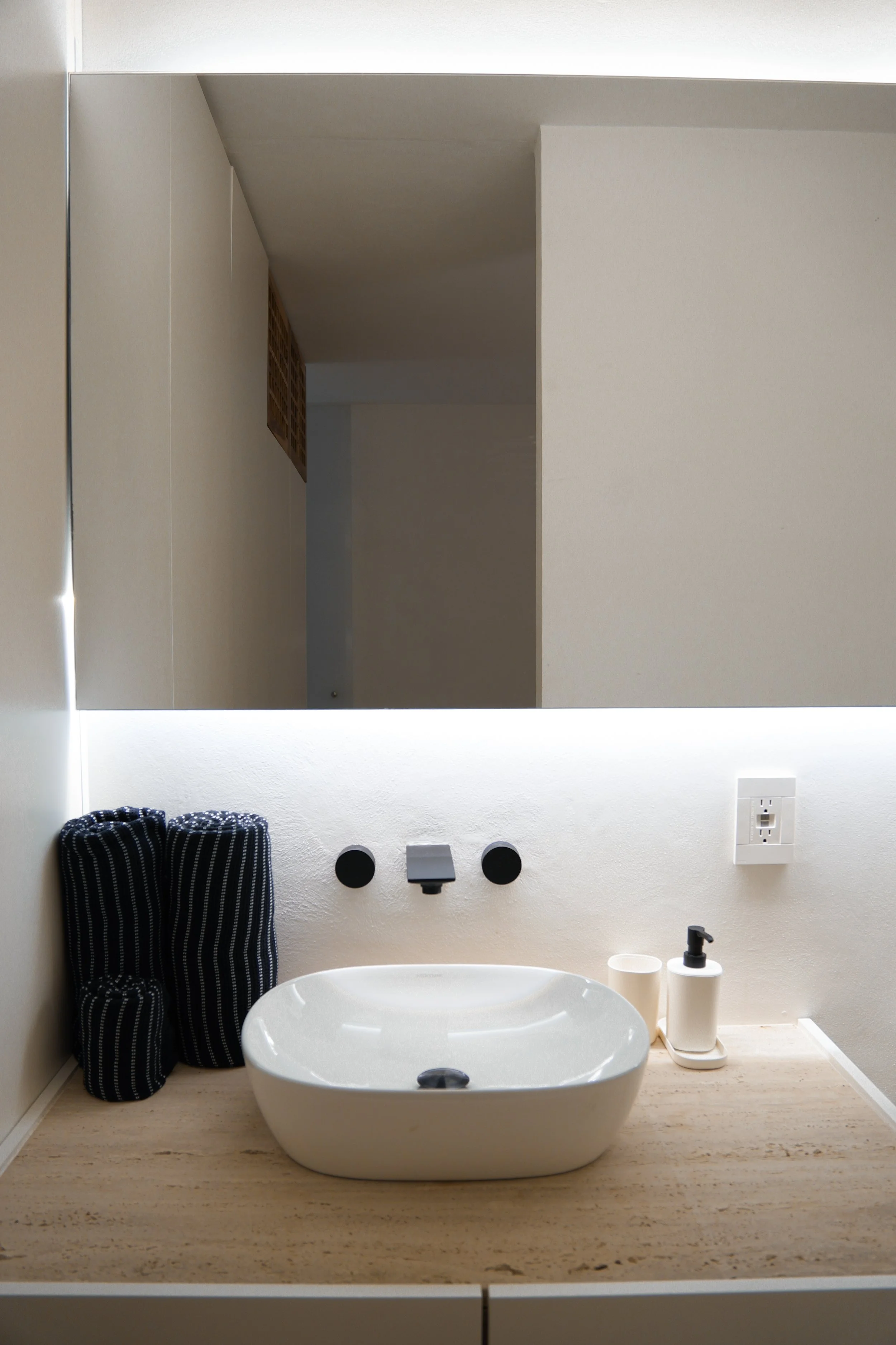 Bathroom vanity area with a wall-mounted mirror and a white vessel sink on a beige countertop. To the left, there is a black and white striped towel hanging. To the right of the sink are a soap dispenser and a small container. The wall behind has a w
