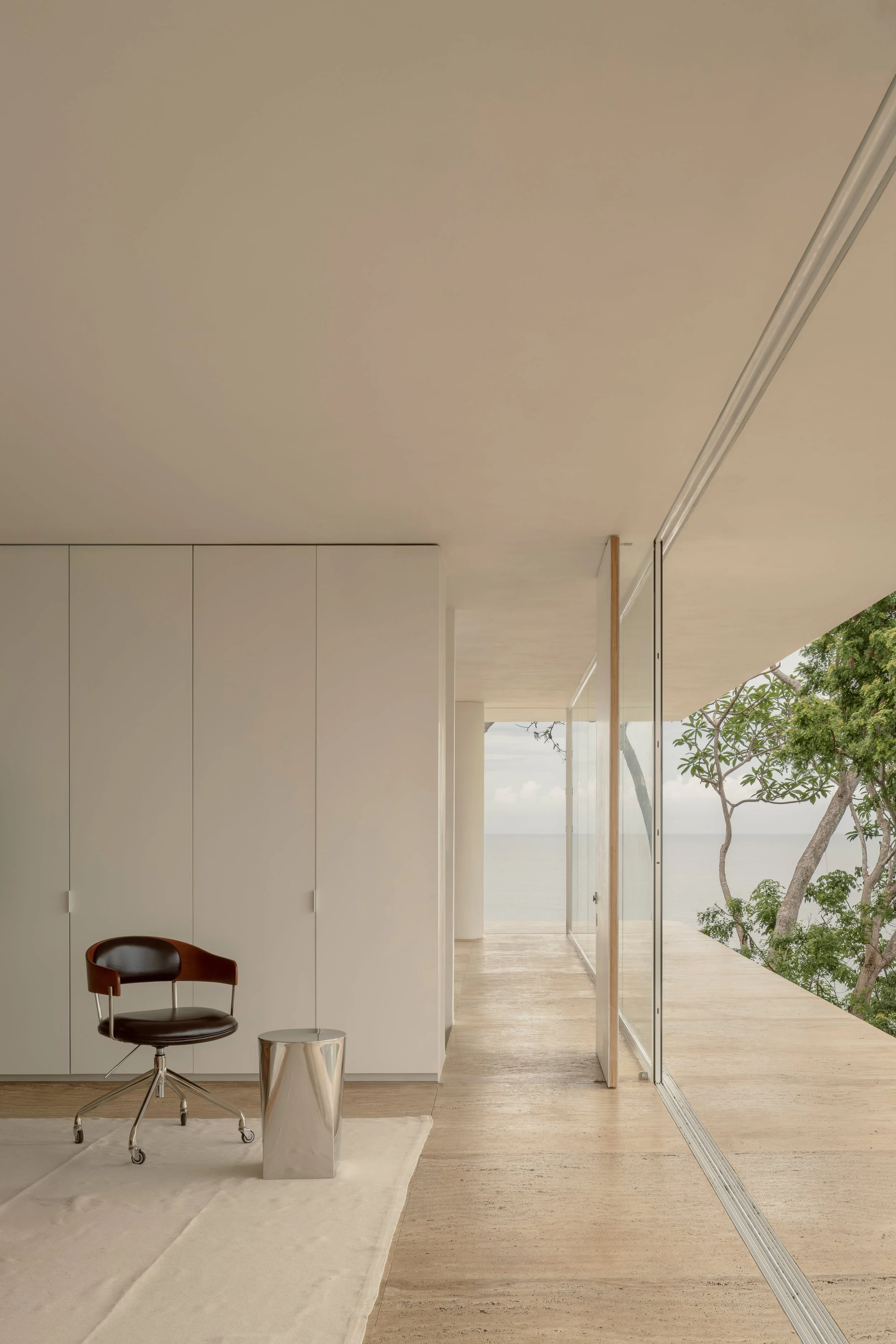Modern interior space with white cabinetry, a black and brown office chair, a metallic side table, a white rug, and large windows showing trees and an ocean view outside.