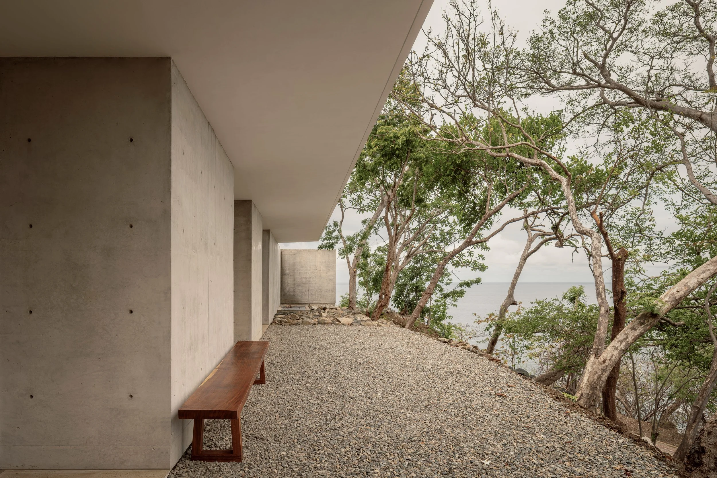 Modern concrete building with a white ceiling, wooden bench, gravel pathway, and trees overlooking the ocean in the distance.