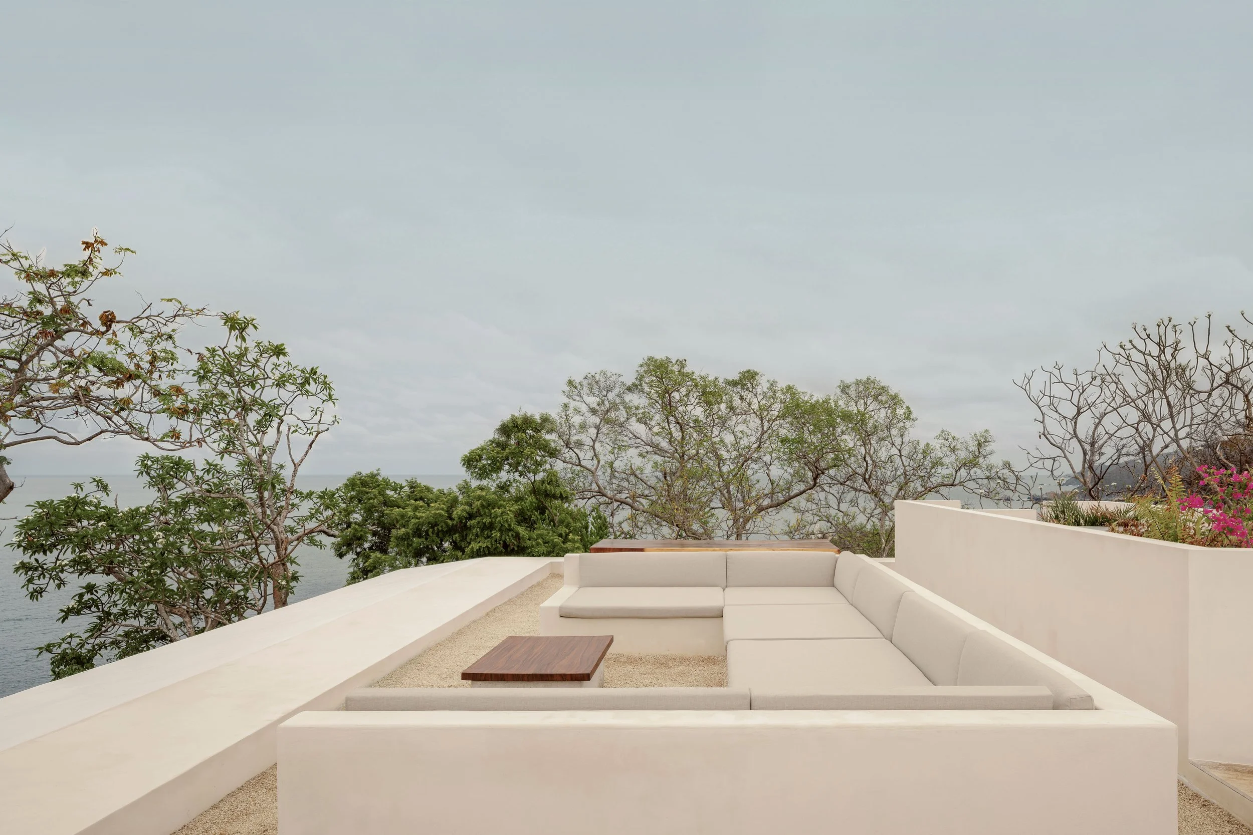 A white outdoor patio with a white sectional sofa and small wooden coffee table, overlooking trees and the water under an overcast sky.