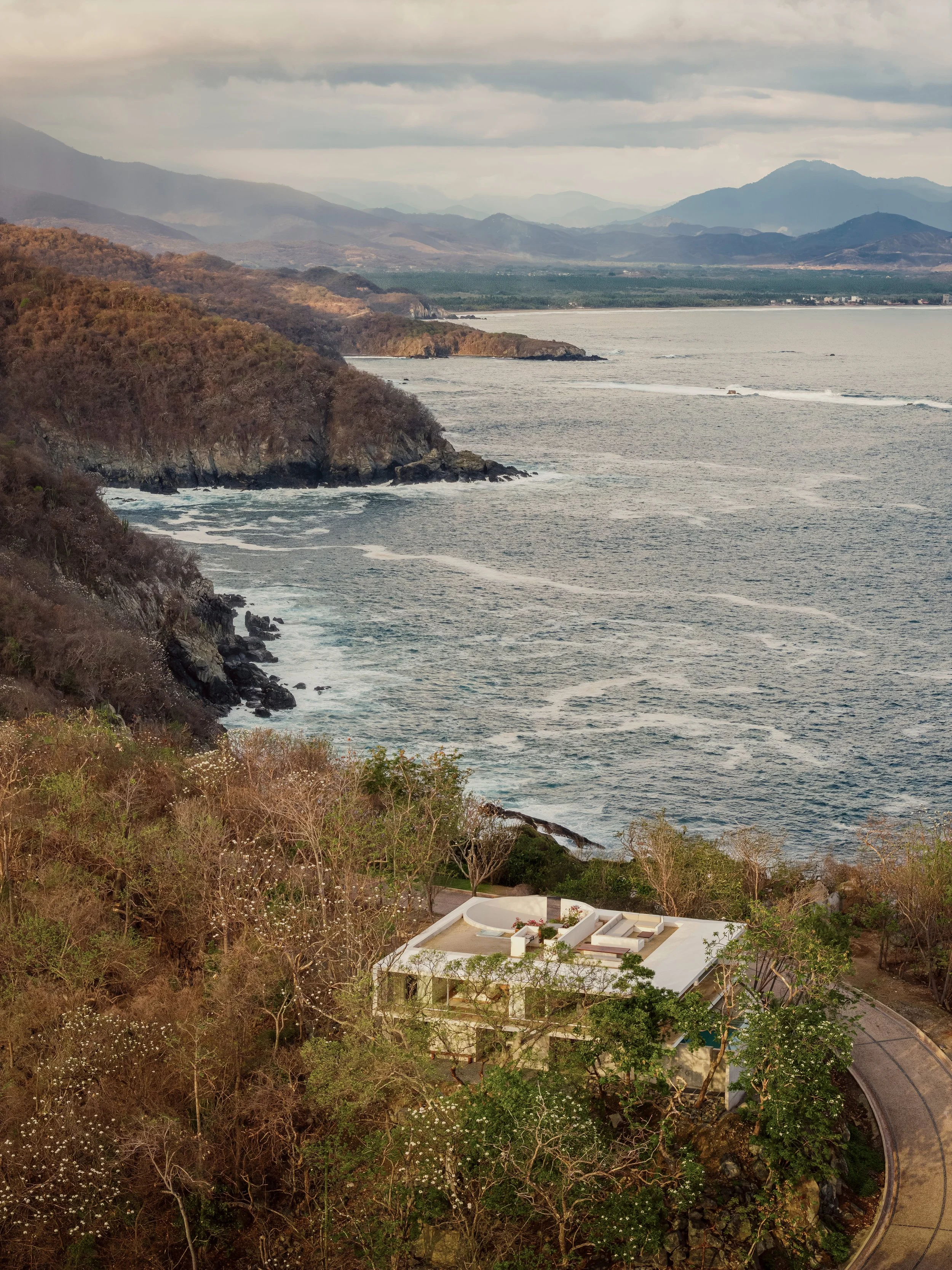 A coastal landscape with rocky cliffs, the ocean, and mountains in the background, featuring a modern white building with a curved roof and surrounding greenery.