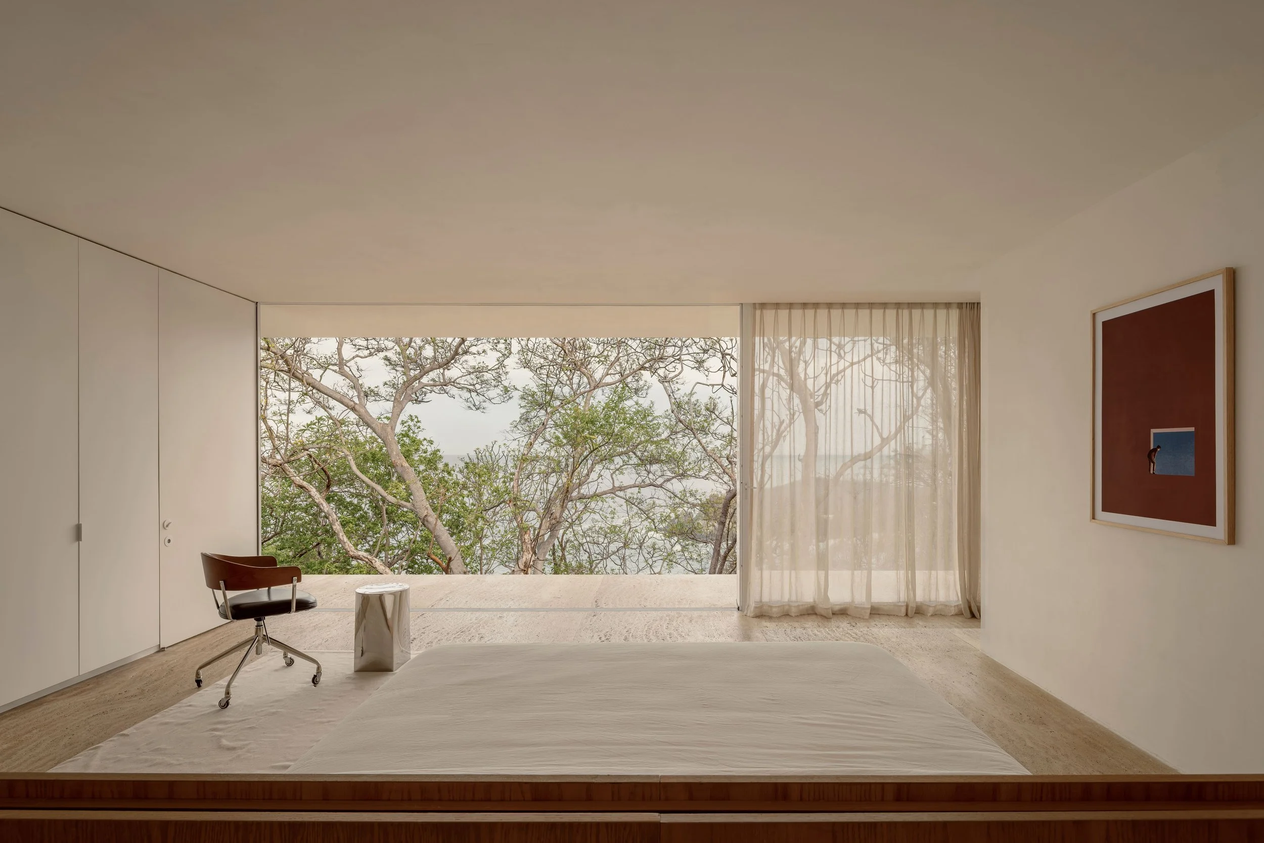 Minimalist bedroom with large window showing tree branches outside, white curtains, framed artwork on the wall, and a simple stool next to a bed.