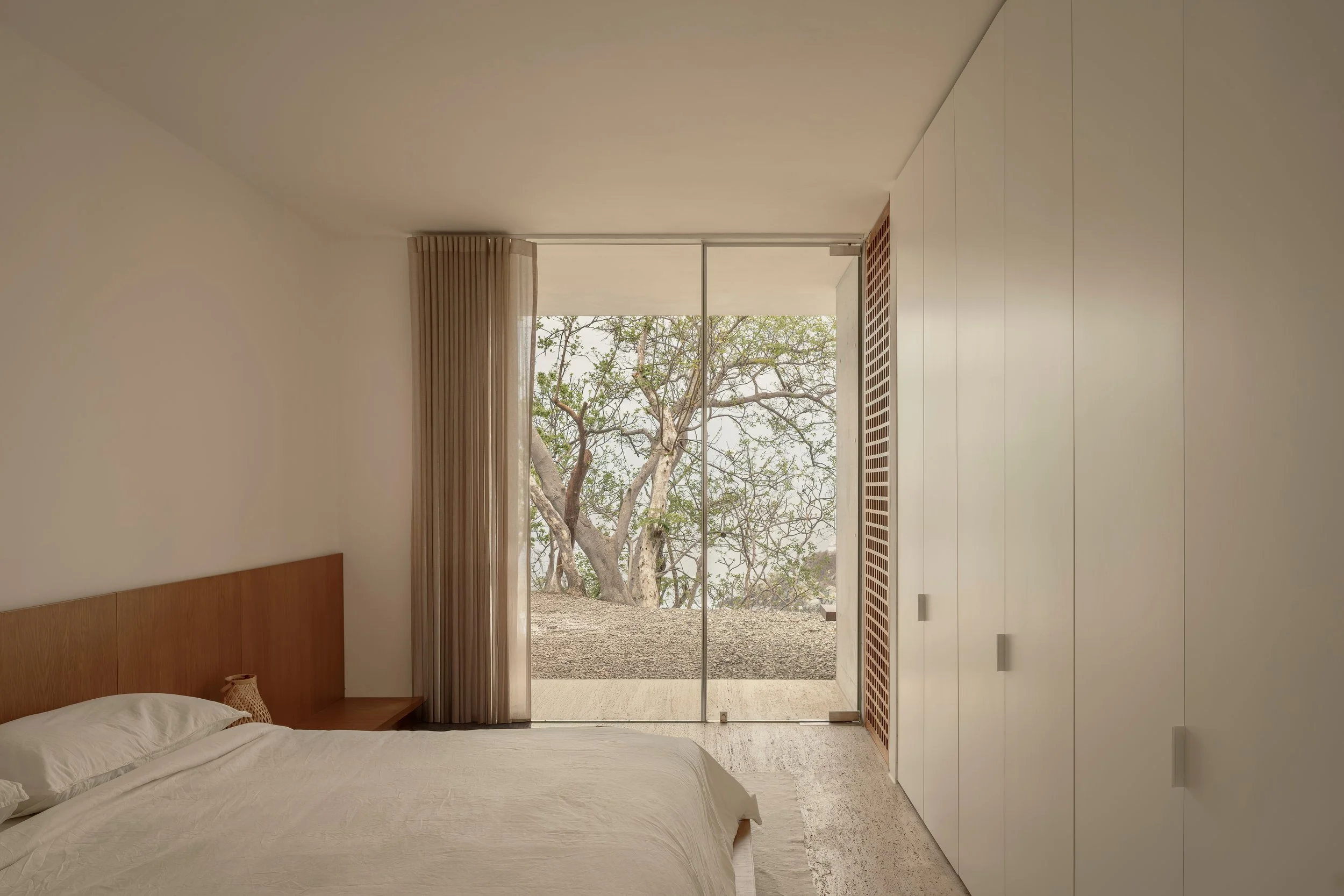 A minimalist bedroom with a large window showing trees outside. The bed has white bedding and a wooden headboard, positioned against the left wall. Beige curtains partially cover the window, and built-in white storage units are on the right. The floo