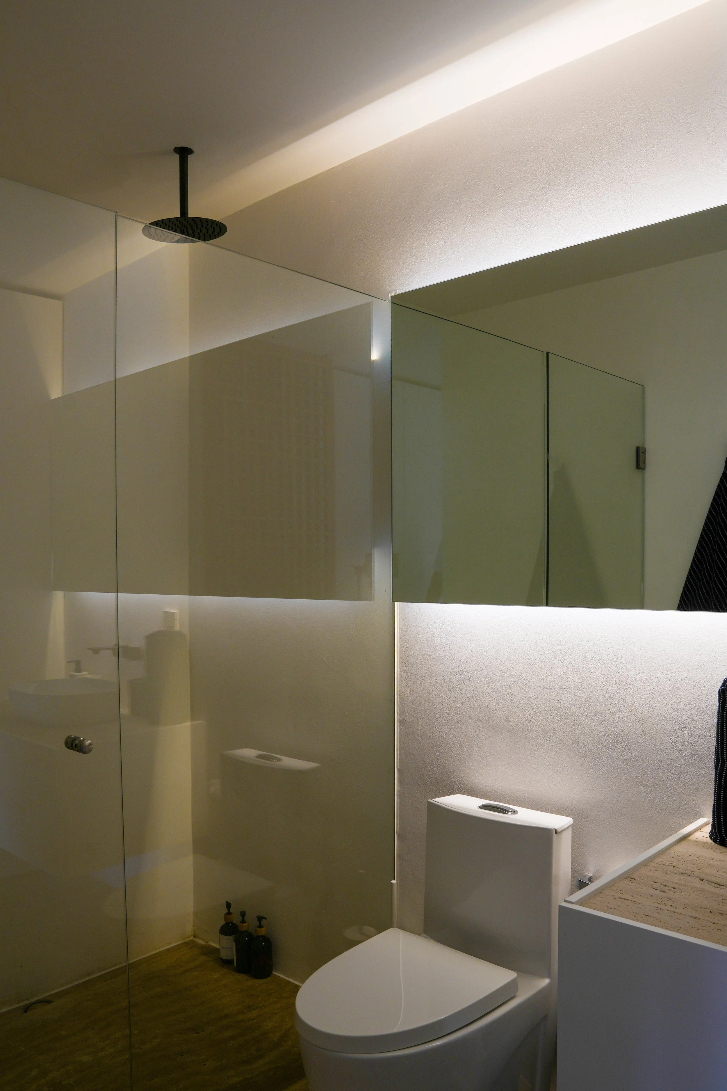 A modern bathroom with a walk-in shower, large mirror, white toilet, and beige walls and flooring. The shower has a black rain showerhead, and some bottles are placed on the floor near the shower.