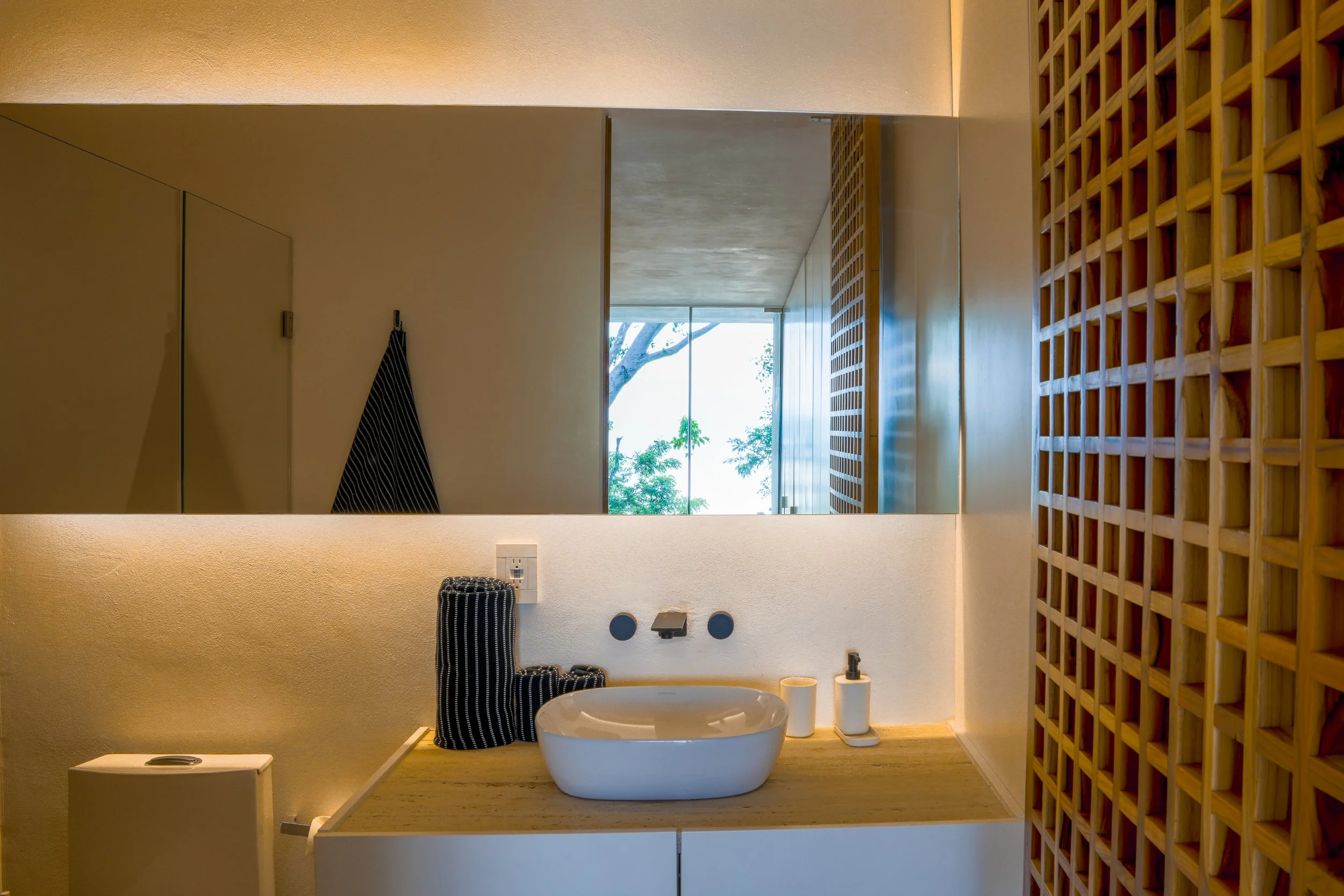 Modern bathroom with a large mirror, wooden lattice wall, white vessel sink, and bathroom accessories on a light wooden countertop. Natural light coming through windows with a view of trees.