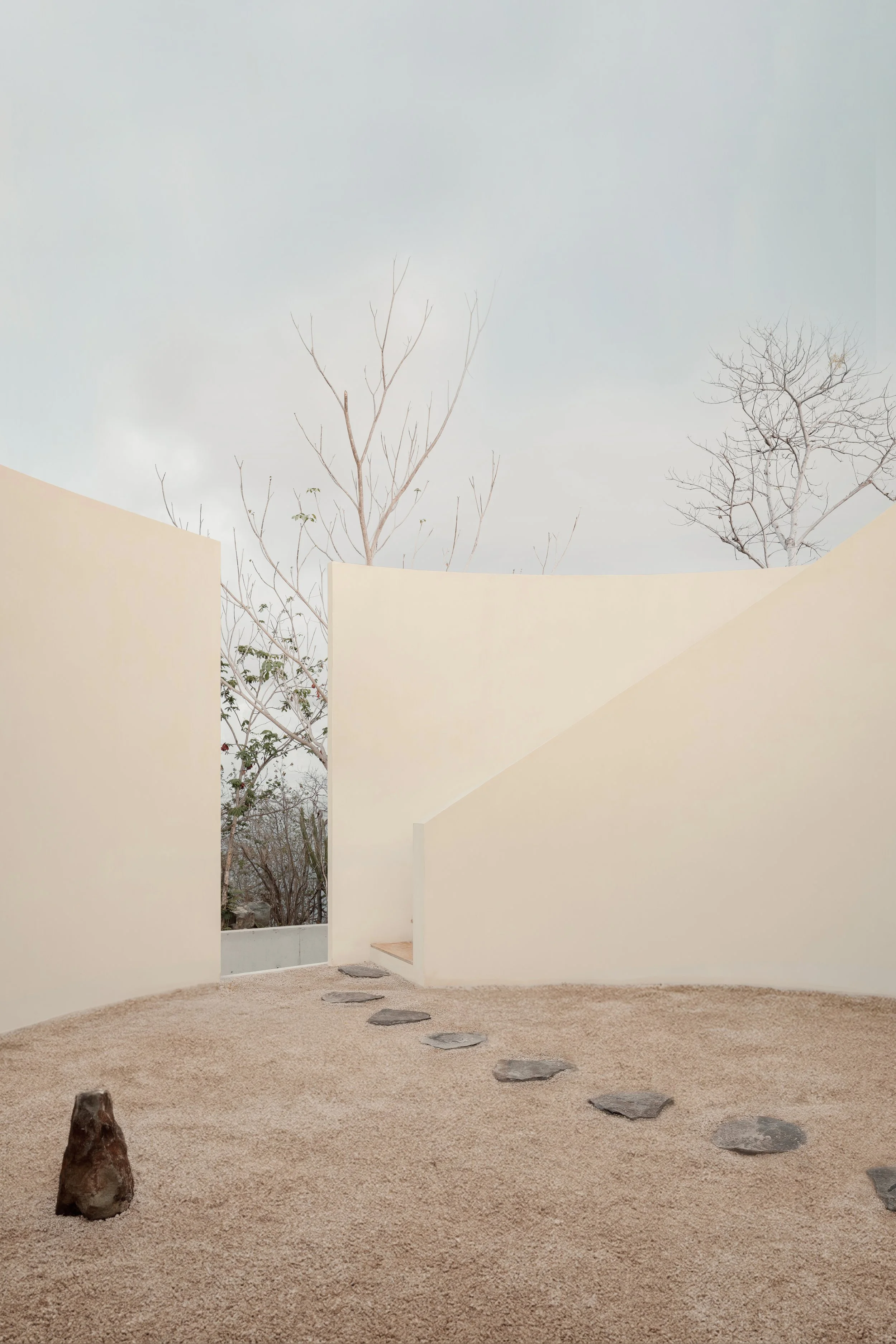 Minimalist courtyard with beige sand ground, stepping stones, cream-colored curved walls, and leafless trees visible through rectangular openings.