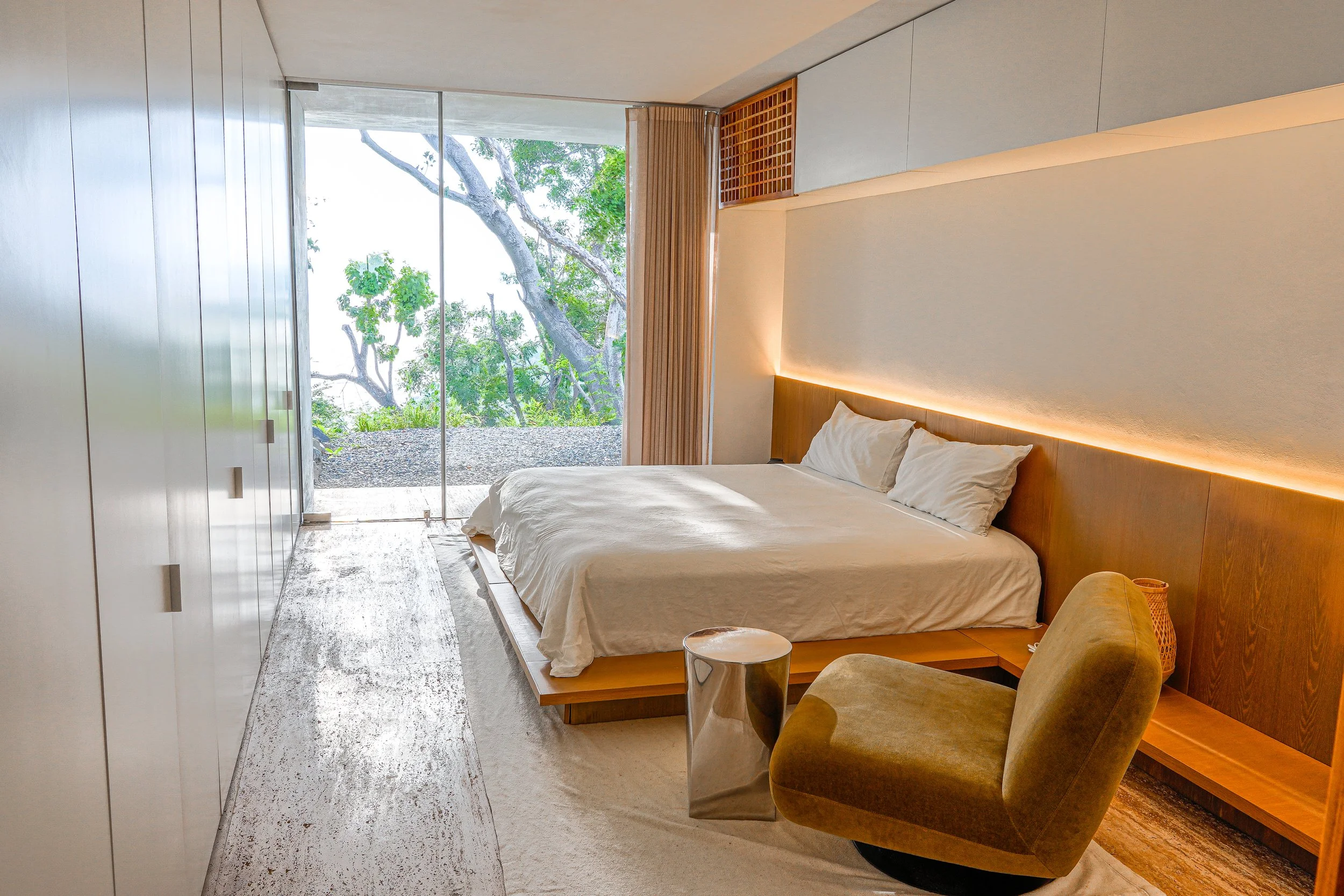 Modern bedroom with a large bed, wood headboard, white bedding, and pillows. A round metallic side table is next to a yellow armchair. Floor-to-ceiling glass window showing trees outside. Wooden panel accents and built-in storage with soft lighting o