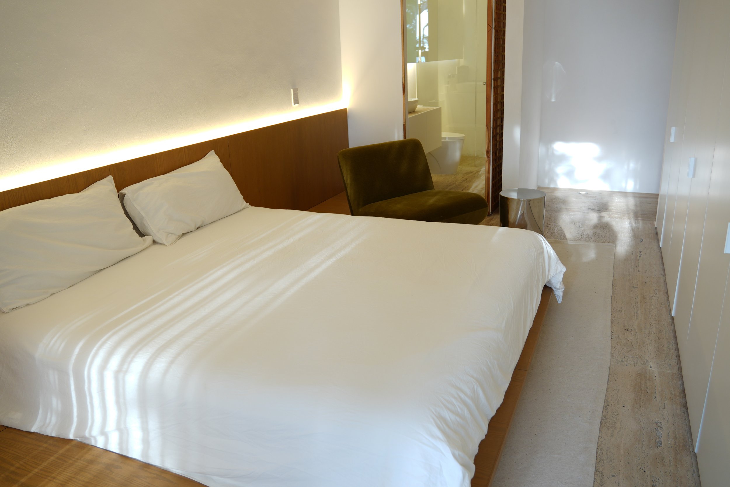 Interior view of a modern hotel room with a bed, a brown chair, small side table, and a bathroom visible through an open door. The room has white walls, a wood headboard, and natural light.
