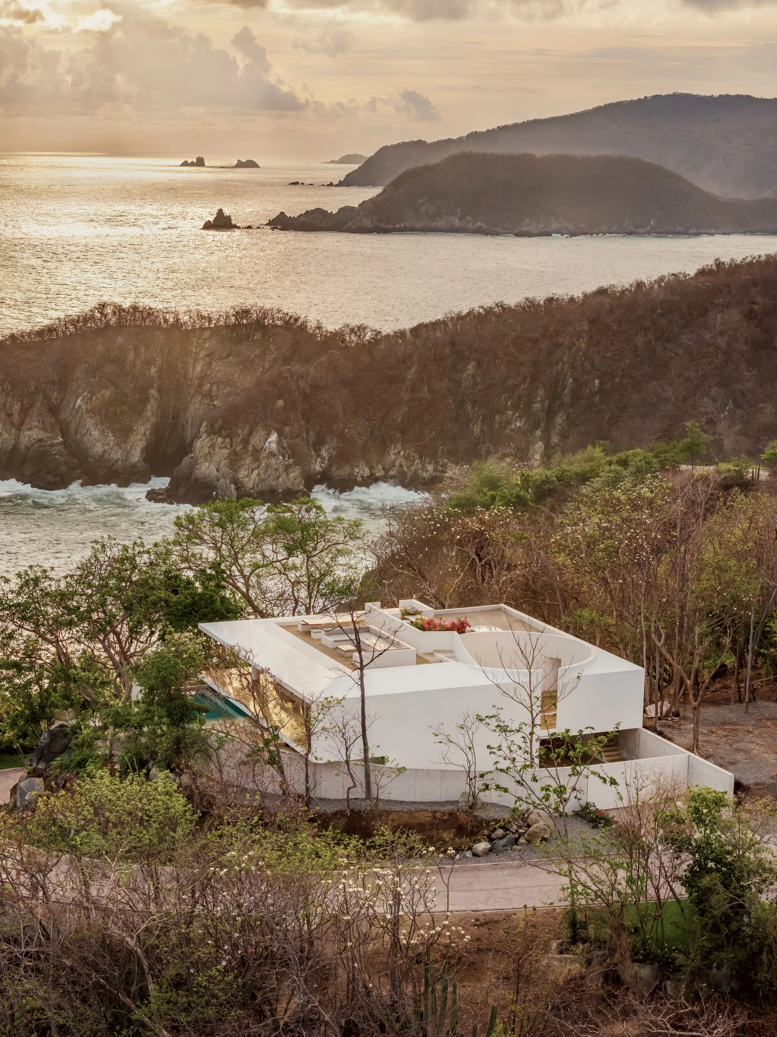 Modern white building with curved walls and rooftop terrace, surrounded by trees, overlooking a rocky coastline with the ocean and hills in the background.
