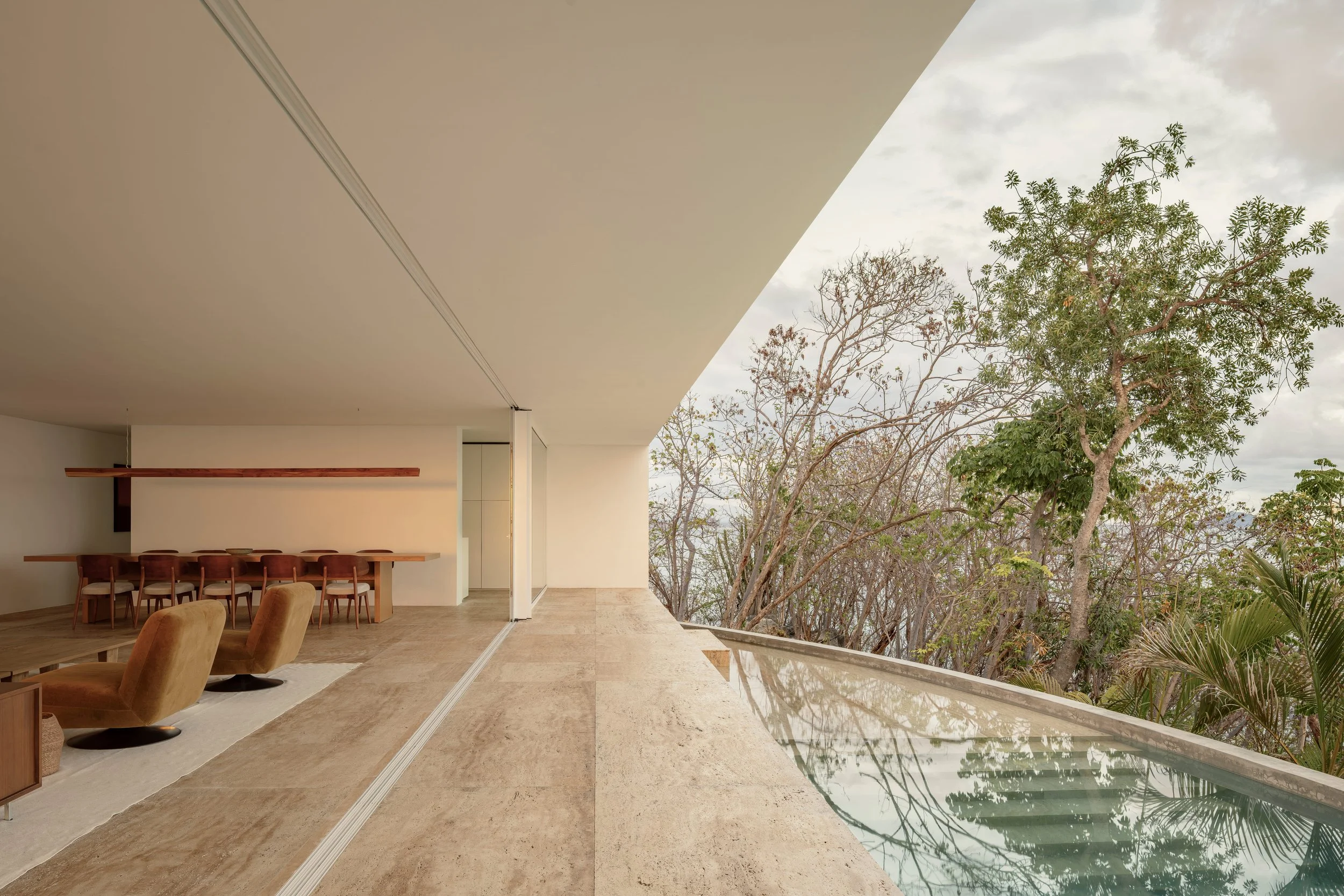 Modern living space with beige marble flooring, beige walls, a long wooden dining table with chairs, and a large sliding glass door opening to an outdoor area with trees and cloudy sky.
