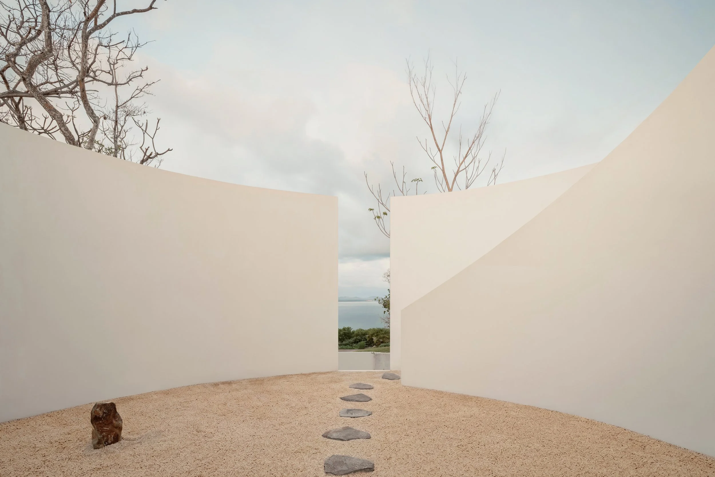 A minimalist outdoor scene with a sandy ground, a large rock, and stepping stones leading to an opening between white walls. Bare trees are visible through the opening against a cloudy sky and distant water in the background.