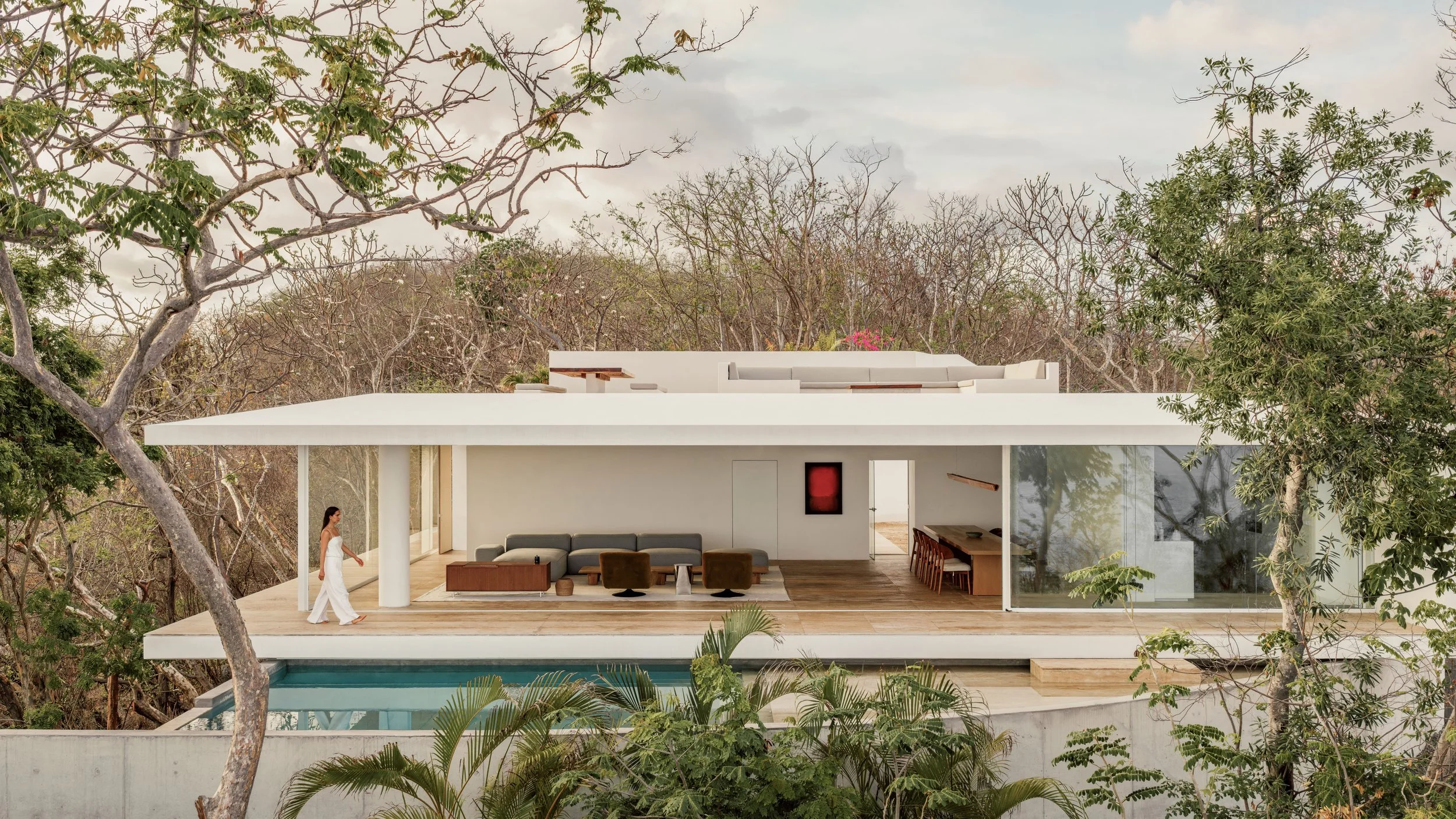 Modern white house with open living space and large glass windows, surrounded by trees, with a swimming pool in front.