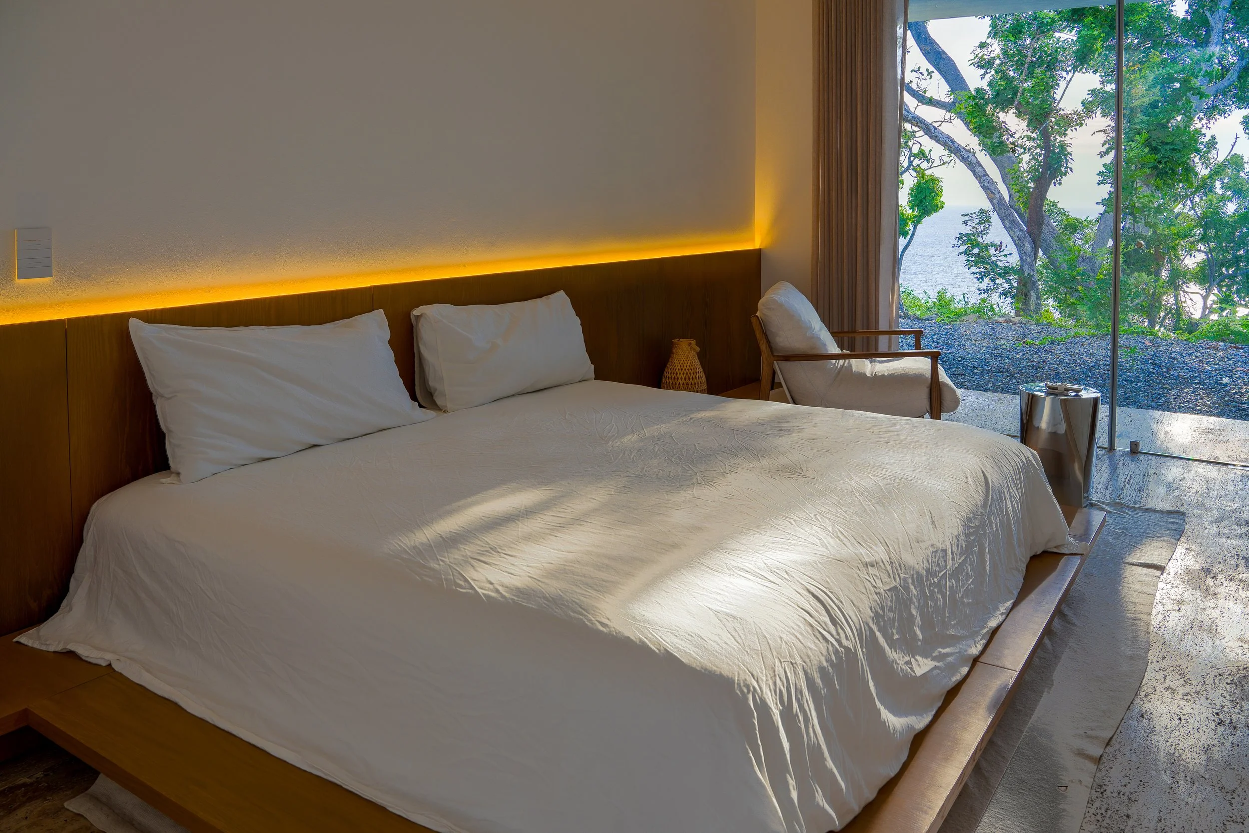 A bedroom with a large bed, white bedding, and pillows. There is a chair and a side table near a large window overlooking trees and a body of water outside.