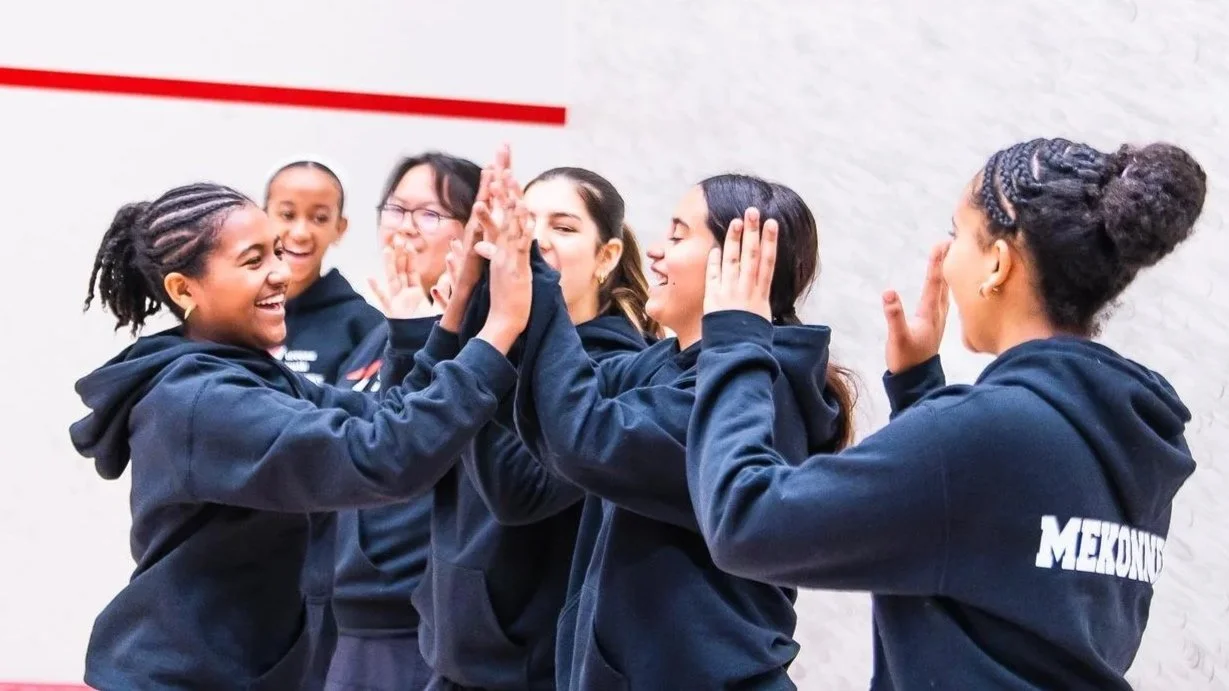 Group of teen girls in dark, team hoodies celebrating with high-fives and smiles inside of a squash court.