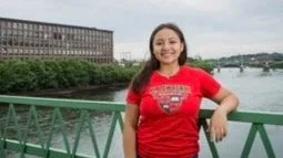 A smiling woman in a red t-shirt with her Lawrence college logo on the front. She is standing on a bridge with green railing and overlooking a river and cityscape. 