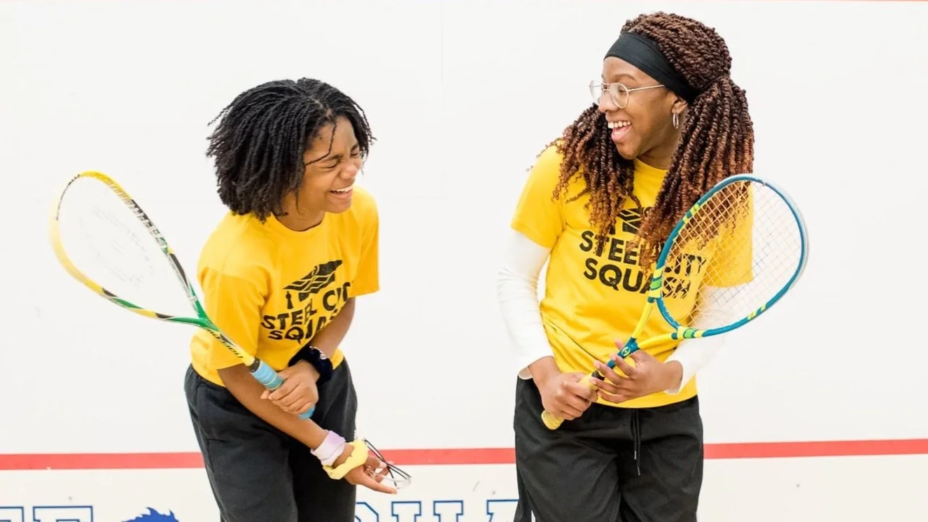 Two teenage girls wearing yellow 'Steel City Squash' t-shirts smiling and laughing while playing squash in an indoor court.