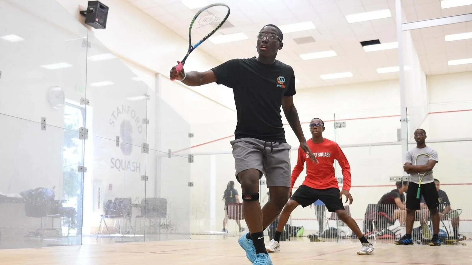Two teenage boys playing squash in an indoor court with a coach in the background. The white, well lit court has a logo on the glass wall reading "Stanford Squash."